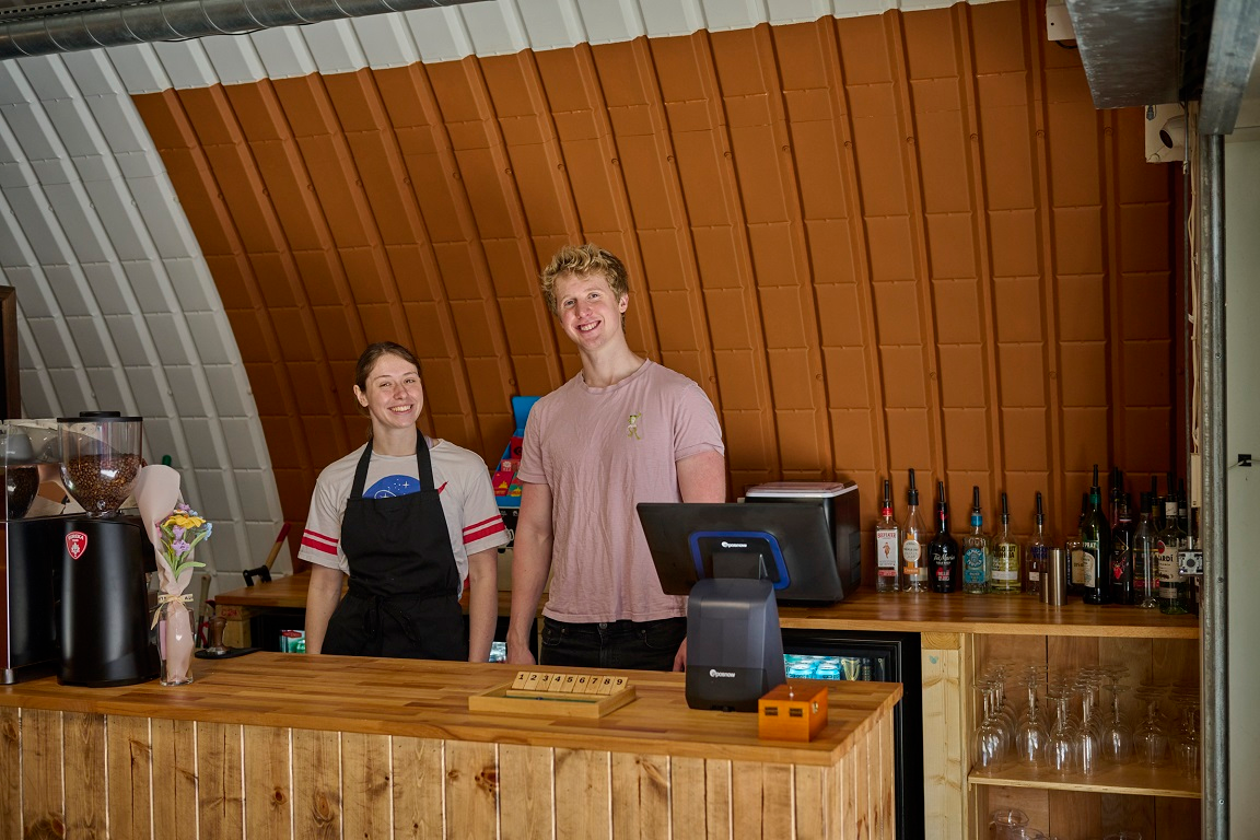 Two smiling people stand behind a wooden bar with a curved, ribbed orange wall in the background.