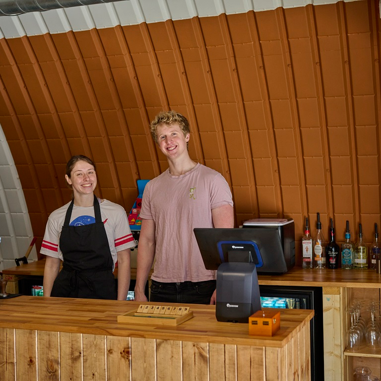 Two smiling people stand behind a wooden bar with a curved, ribbed orange wall in the background.