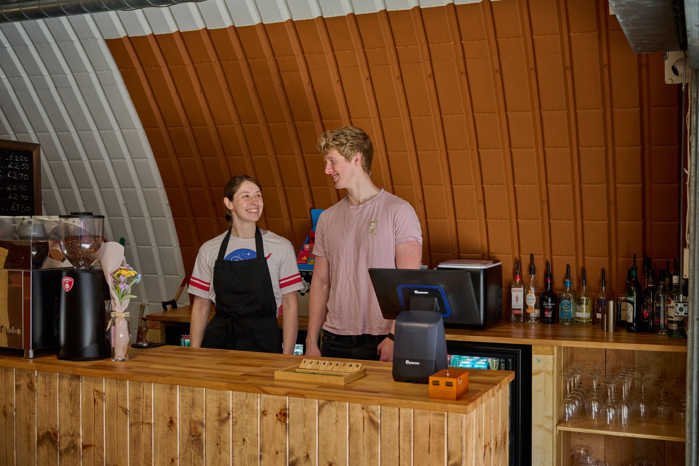 A smiling woman in an apron and a man chat behind a wooden bar.