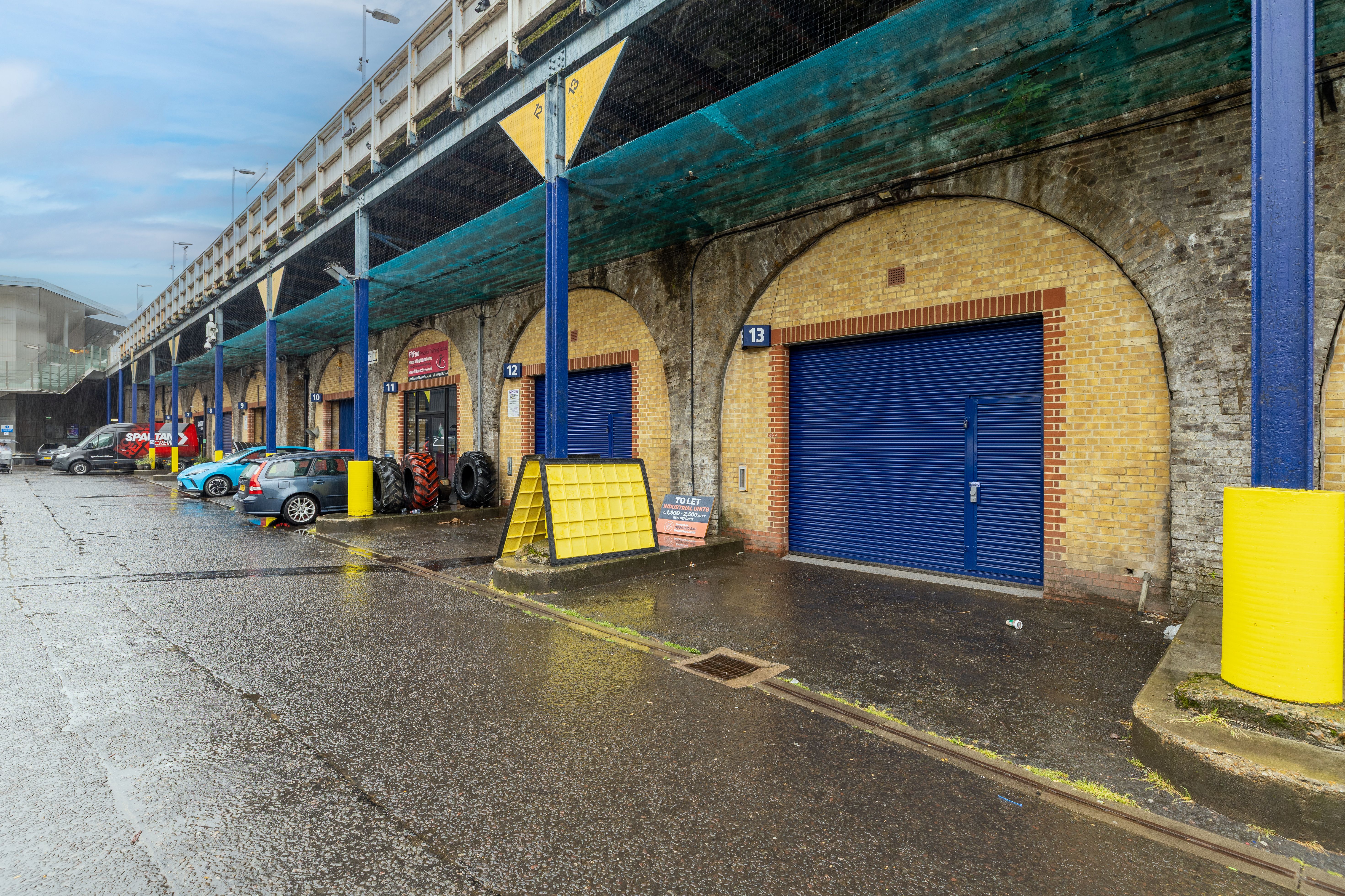 A row of brick archways with blue doors under an elevated structure, with parked cars and wet pavement.