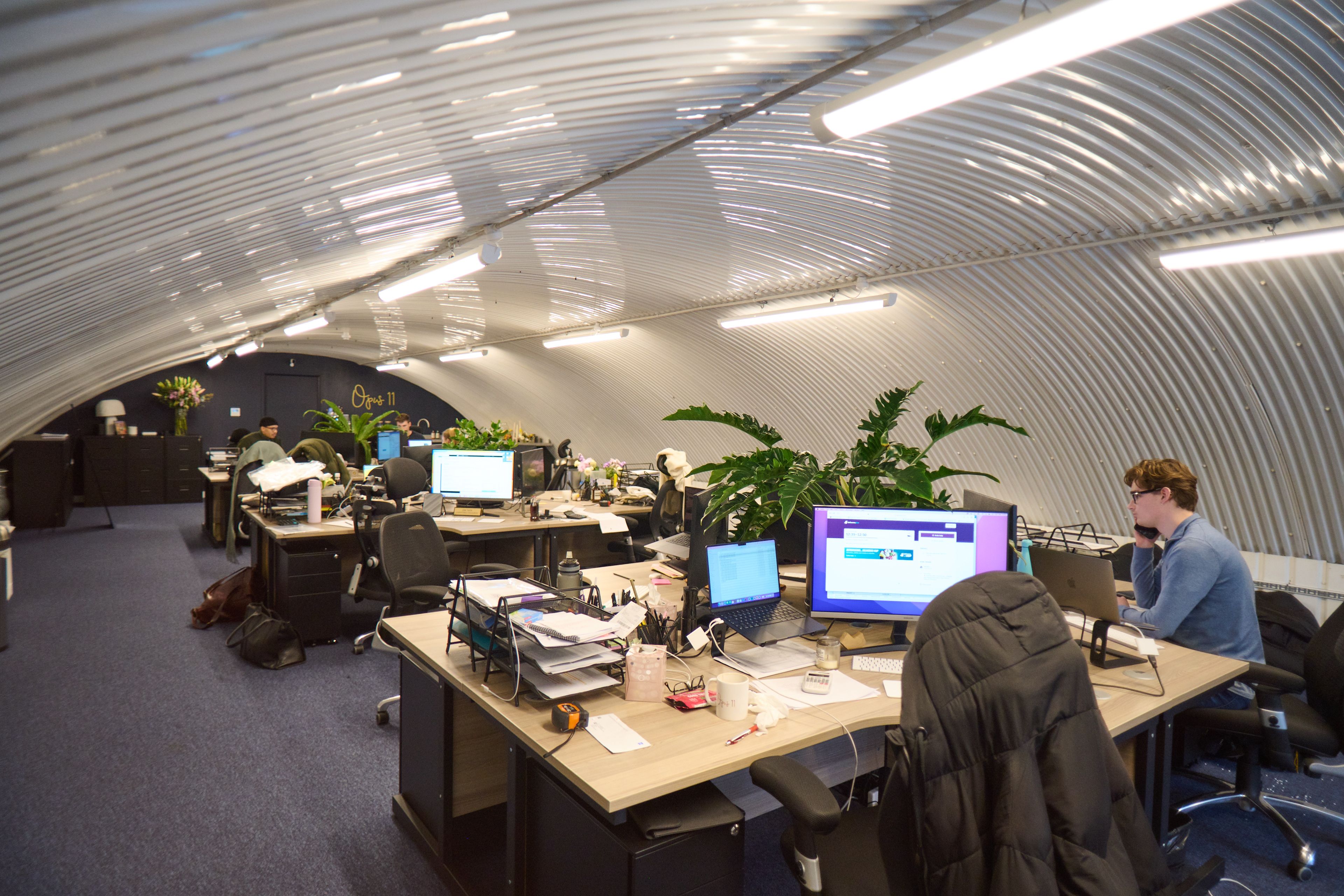 An open-plan office with an arched, corrugated metal ceiling, featuring desks with computers and people working.