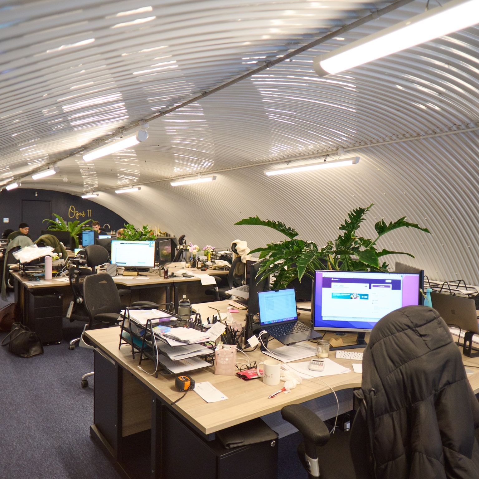 An open-plan office with an arched, corrugated metal ceiling, featuring desks with computers and people working.