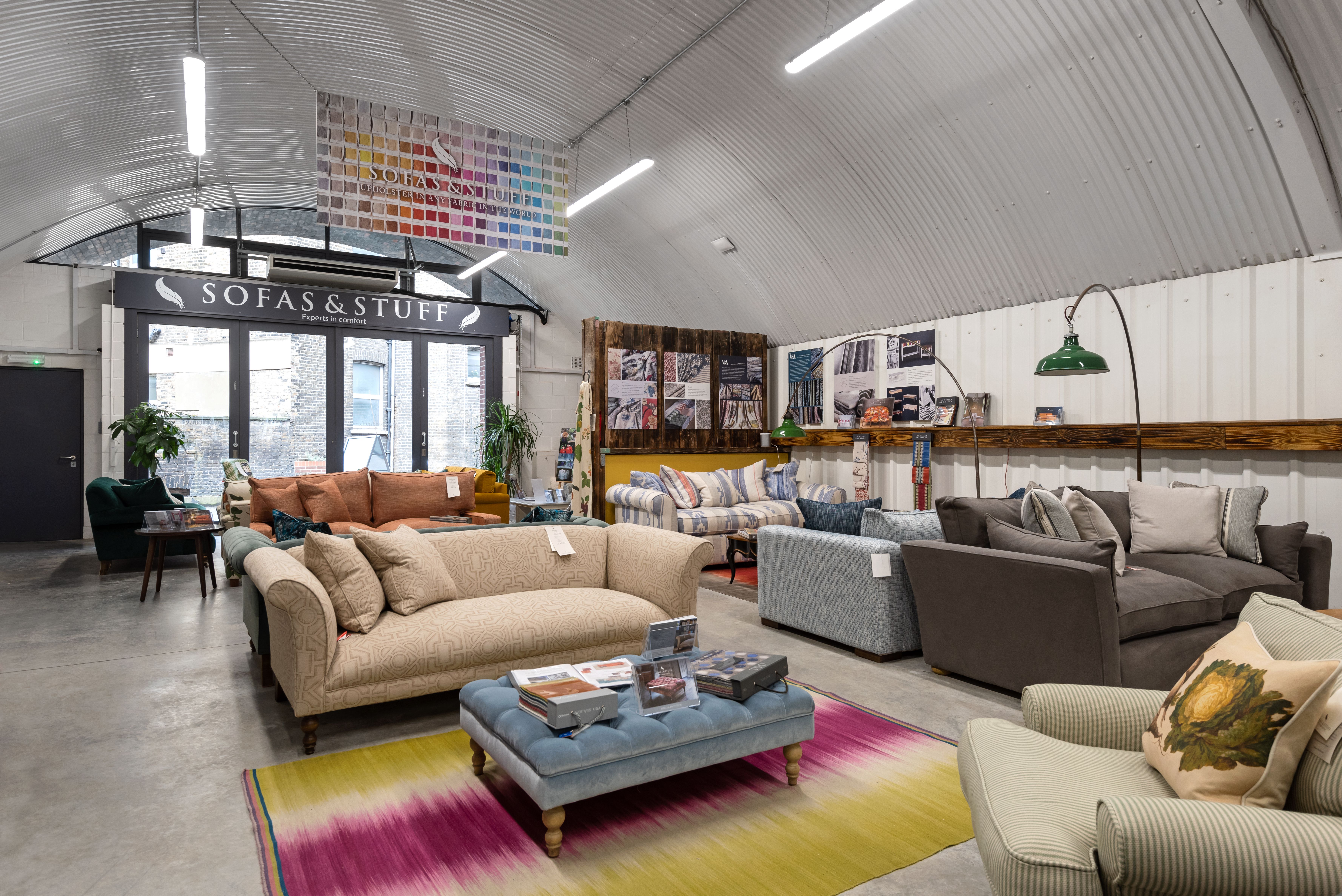Interior of a "Sofas & Stuff" showroom displaying various sofas and armchairs, with a colorful rug and an industrial corrugated ceiling.