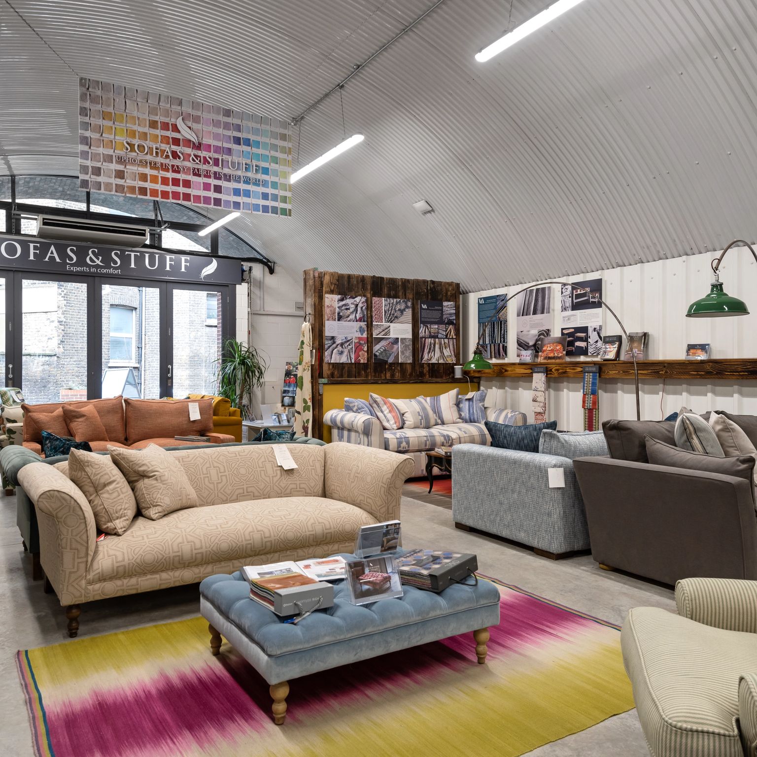 Interior of a "Sofas & Stuff" showroom displaying various sofas and armchairs, with a colorful rug and an industrial corrugated ceiling.