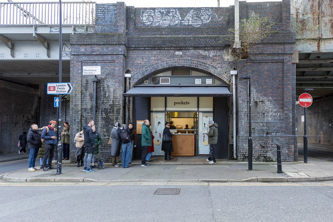 A queue of people waiting outside the 'Pockets' shop, built into a brick railway arch.