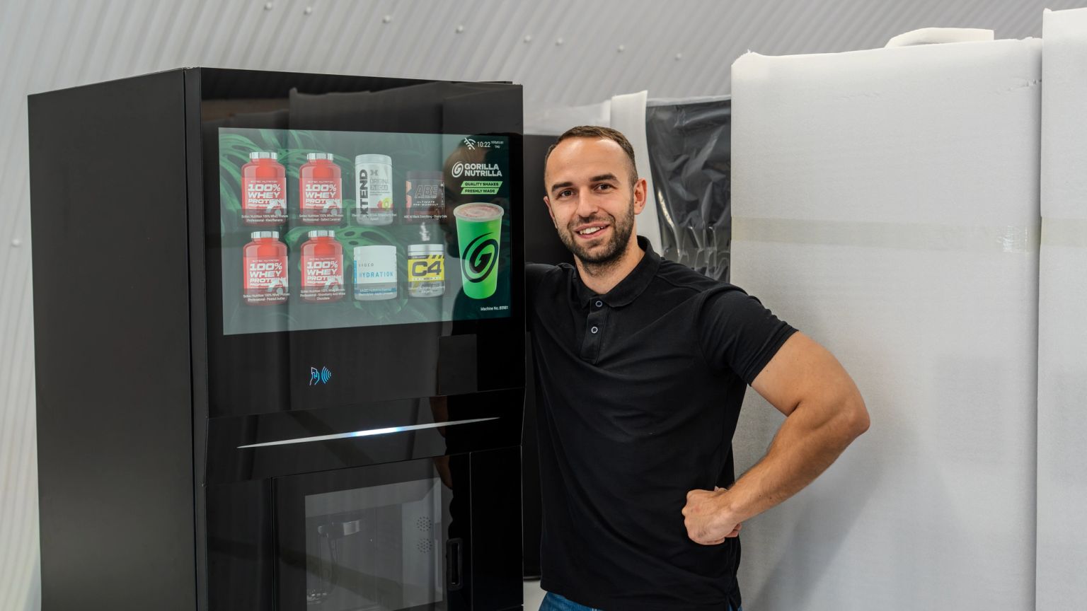 A smiling man stands next to a modern black vending machine displaying fitness supplements.