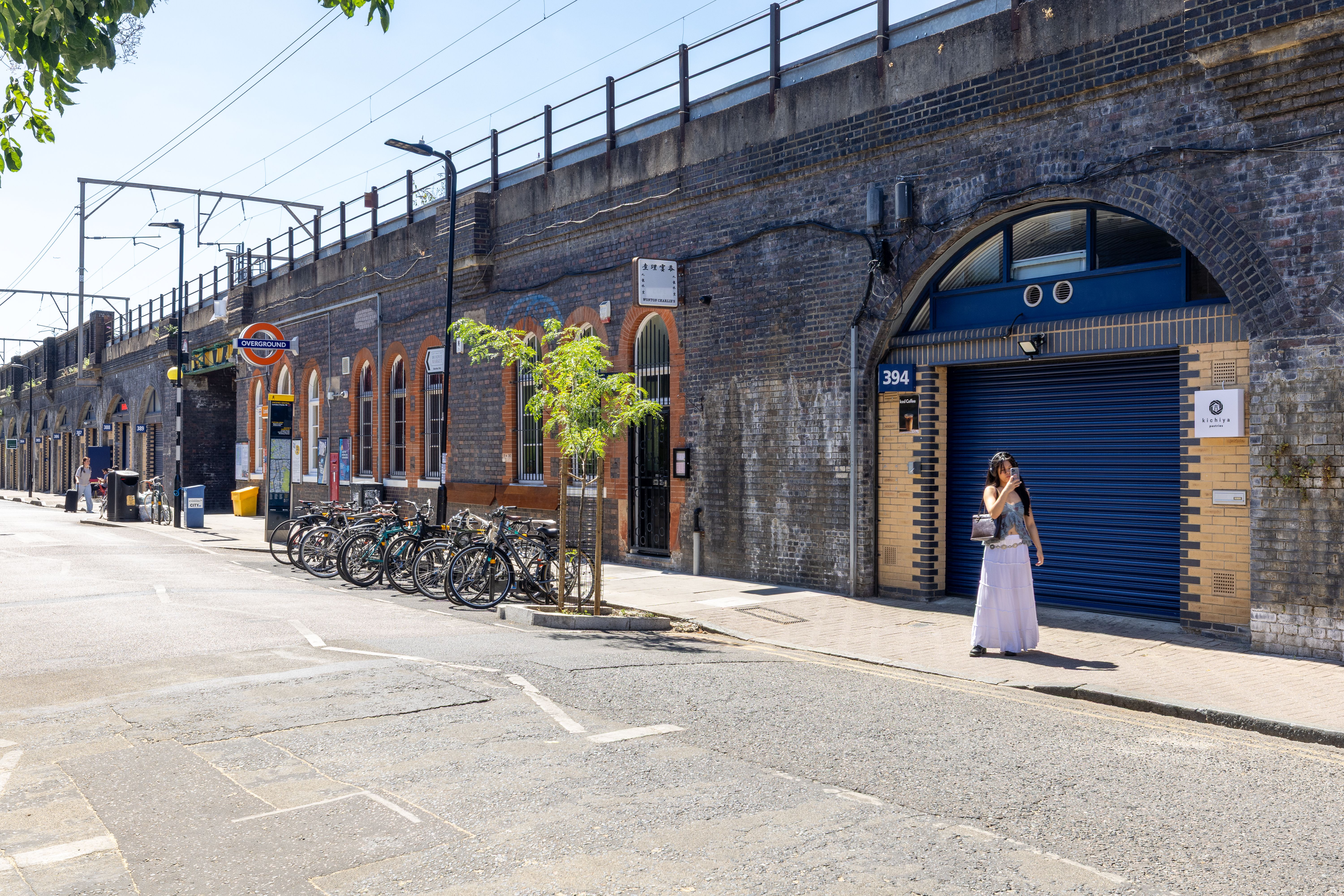A sunny street alongside a long brick railway viaduct with businesses in its arches, a London Underground station sign, parked bicycles, and a woman on the sidewalk.