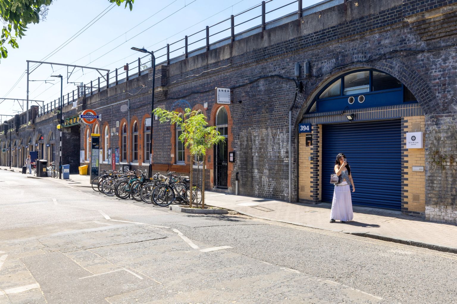 A sunny street alongside a long brick railway viaduct with businesses in its arches, a London Underground station sign, parked bicycles, and a woman on the sidewalk.