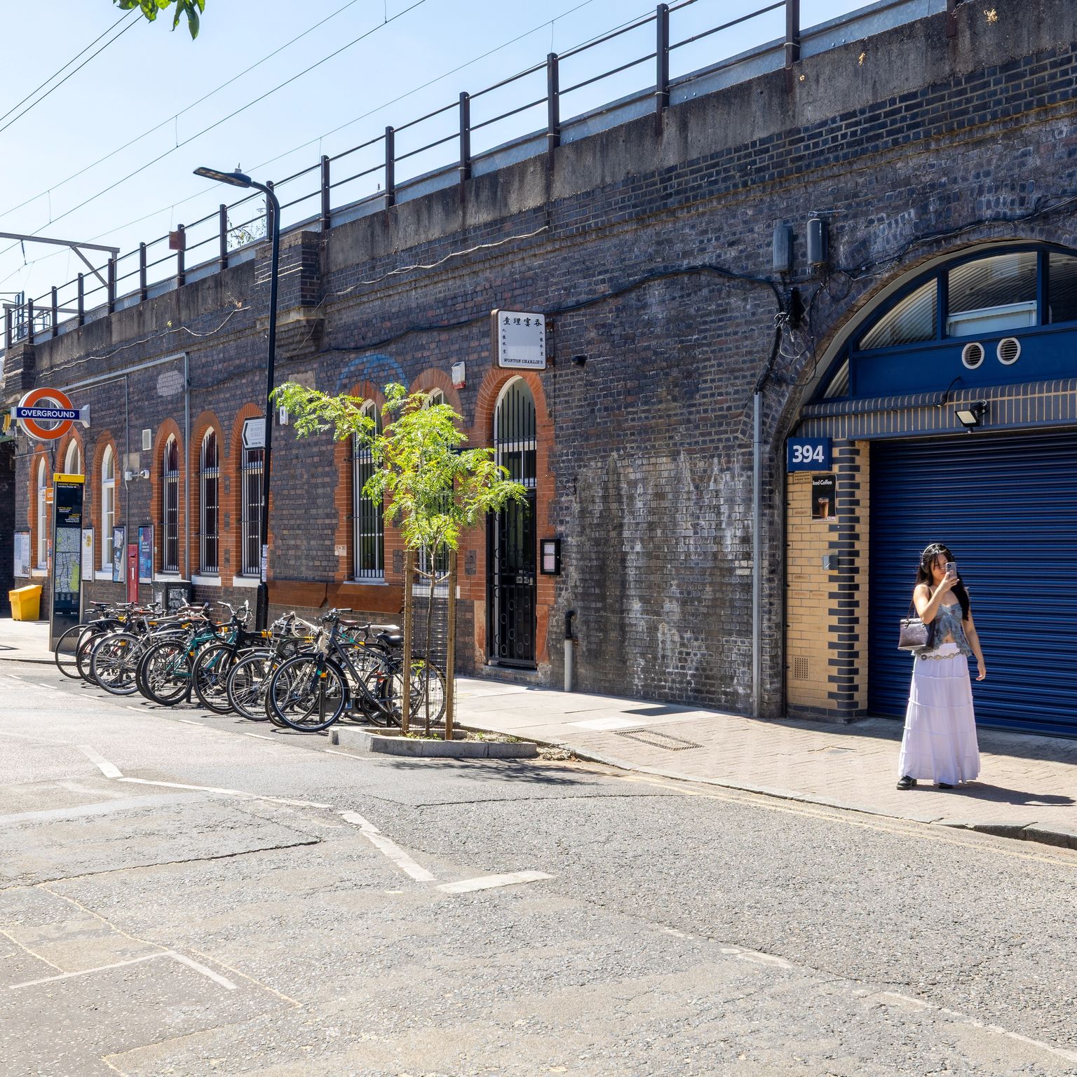 A sunny street alongside a long brick railway viaduct with businesses in its arches, a London Underground station sign, parked bicycles, and a woman on the sidewalk.