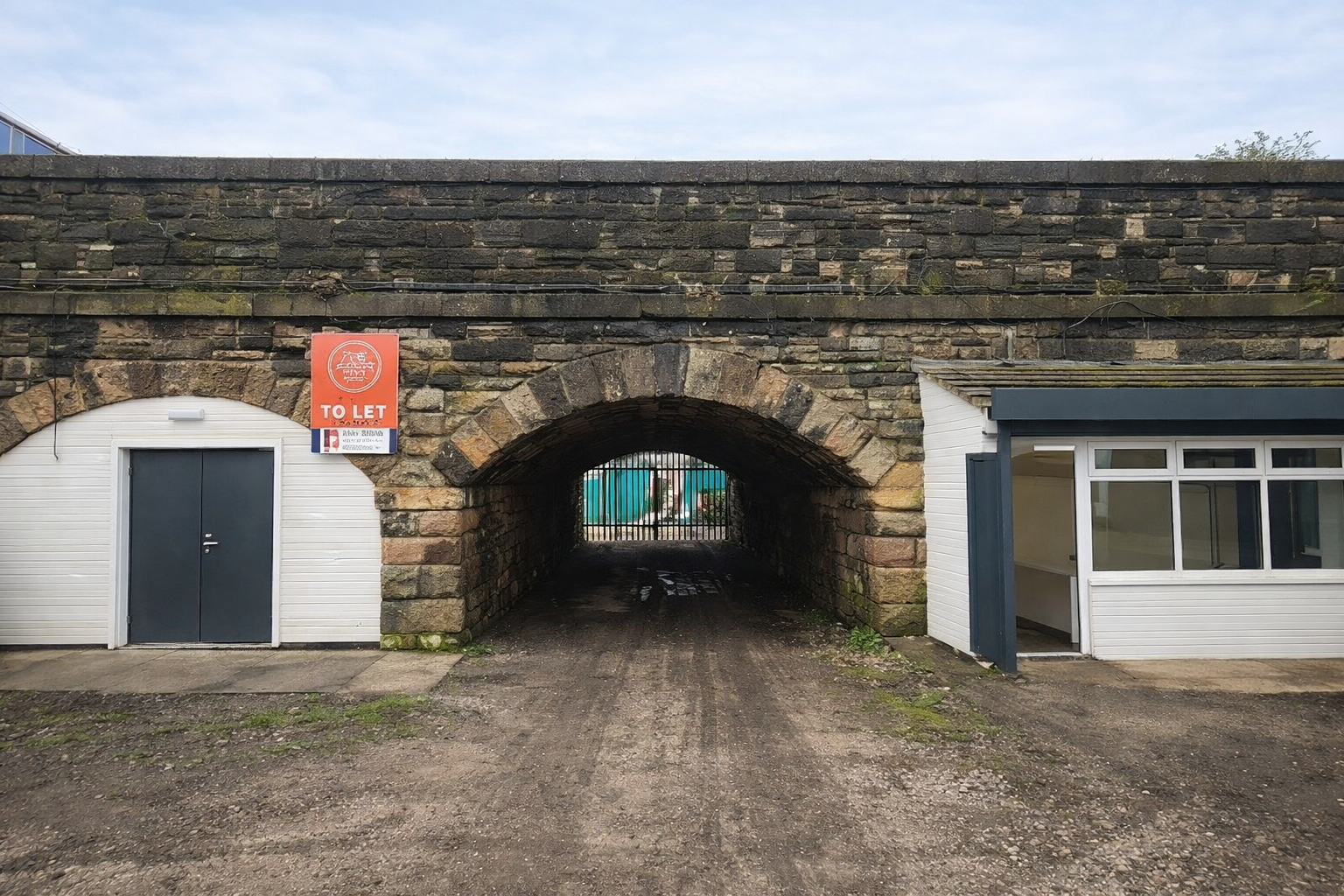 A stone archway over a muddy path, flanked by two small white buildings, one with a "To Let" sign.
