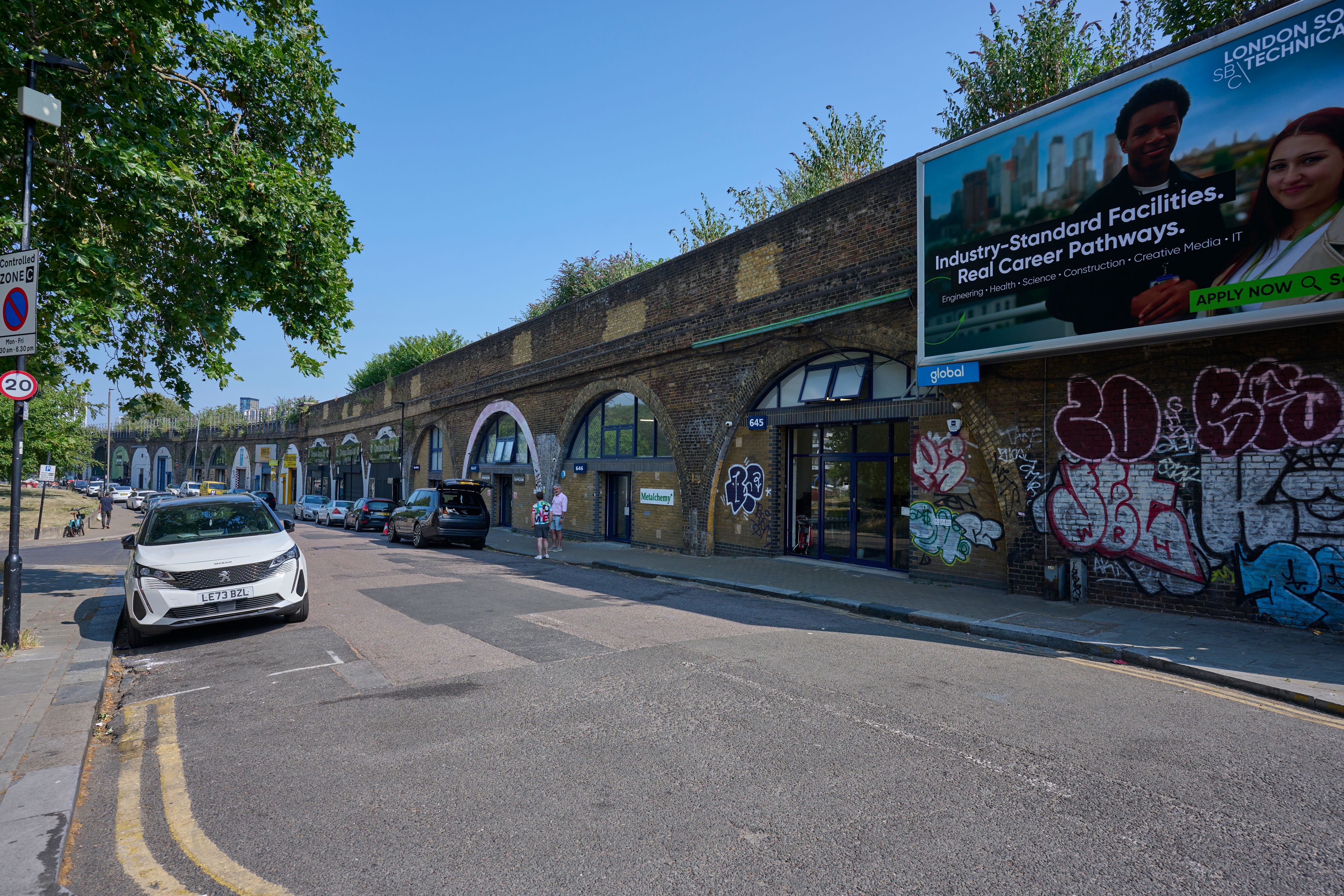 A street runs alongside a long brick viaduct with arched businesses and heavy graffiti, under a clear blue sky.