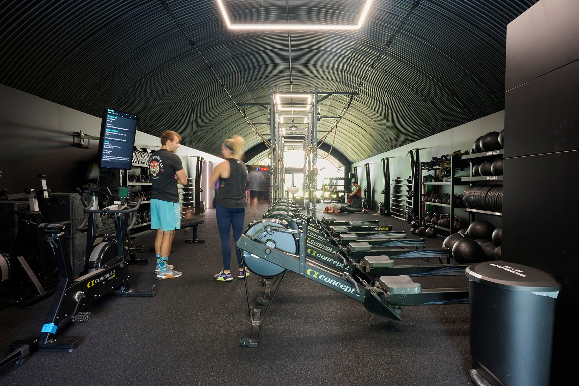 Modern gym interior with a black corrugated arched ceiling, rows of rowing machines, weights, and two people talking.