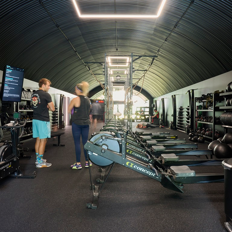 Modern gym interior with a black corrugated arched ceiling, rows of rowing machines, weights, and two people talking.