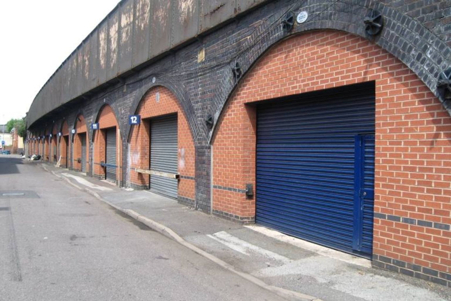 A row of brick archways under a viaduct, with multiple grey and one blue roll-down doors.