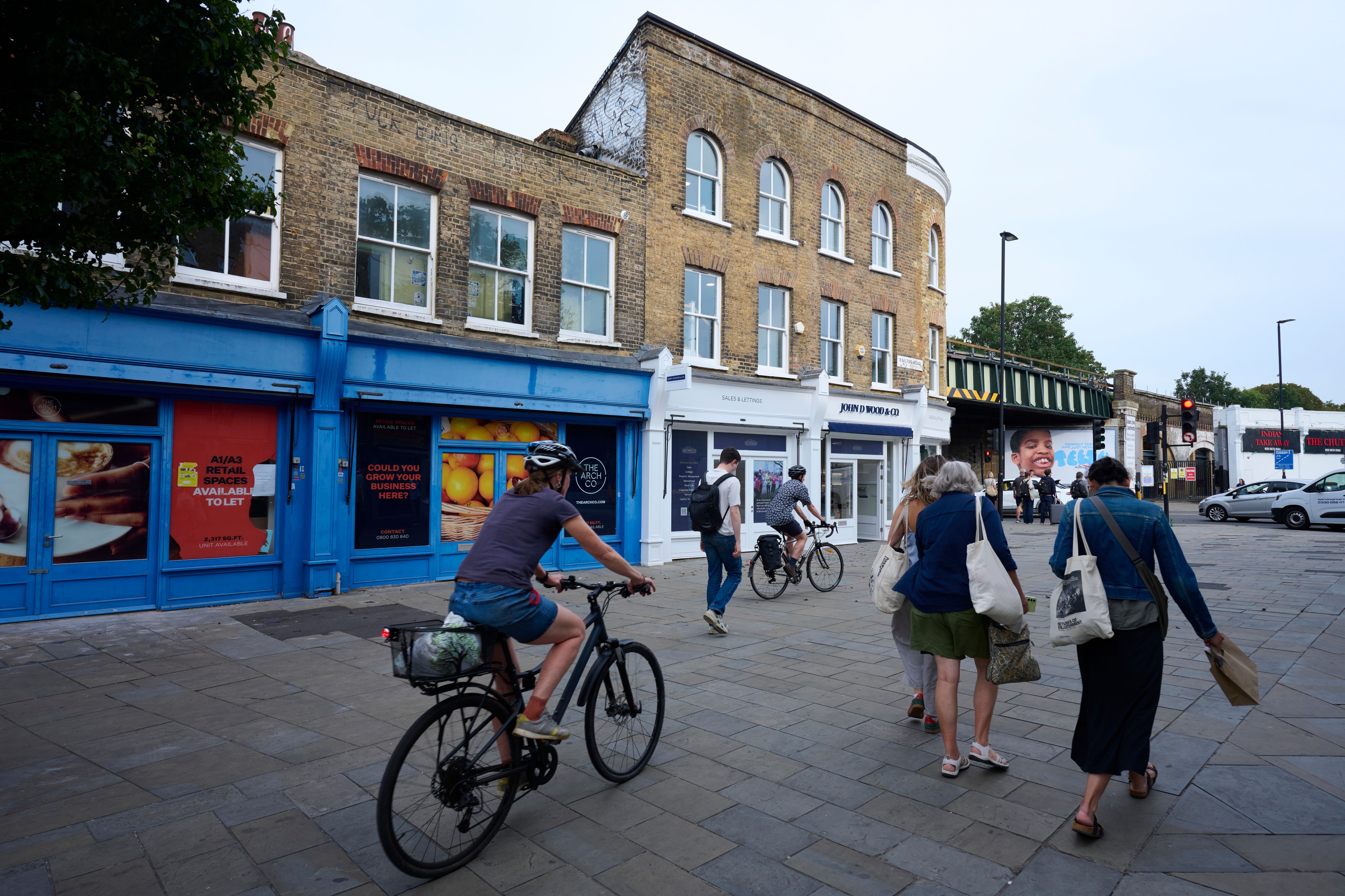 Urban street scene with people walking and cycling past brick buildings and shops, with a railway bridge in the background.