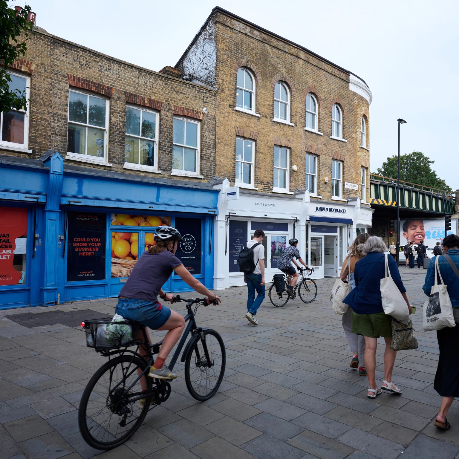 Urban street scene with people walking and cycling past brick buildings and shops, with a railway bridge in the background.