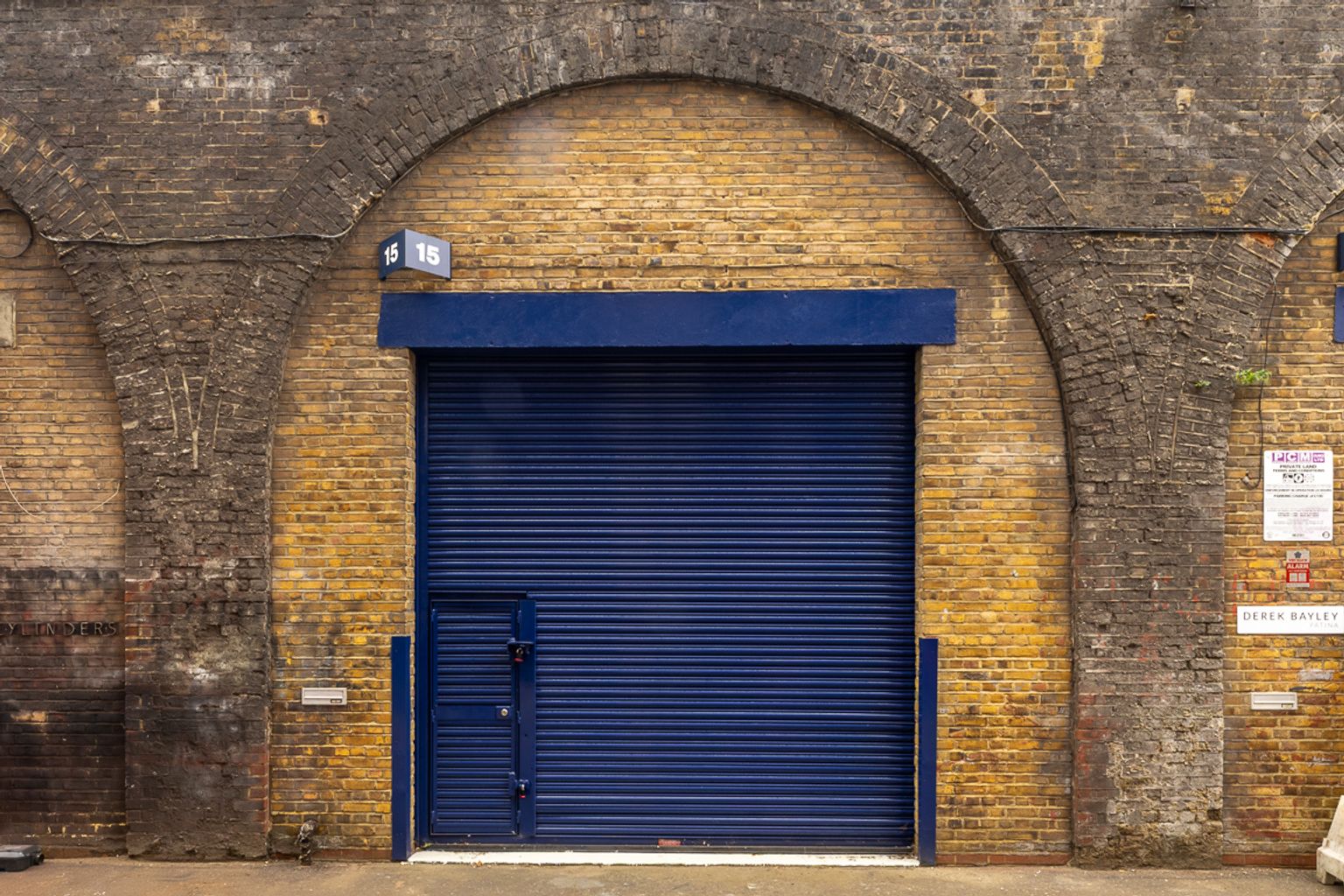 A large dark blue roll-down door with a small pedestrian door, set into a weathered brick archway with the number 15 above.