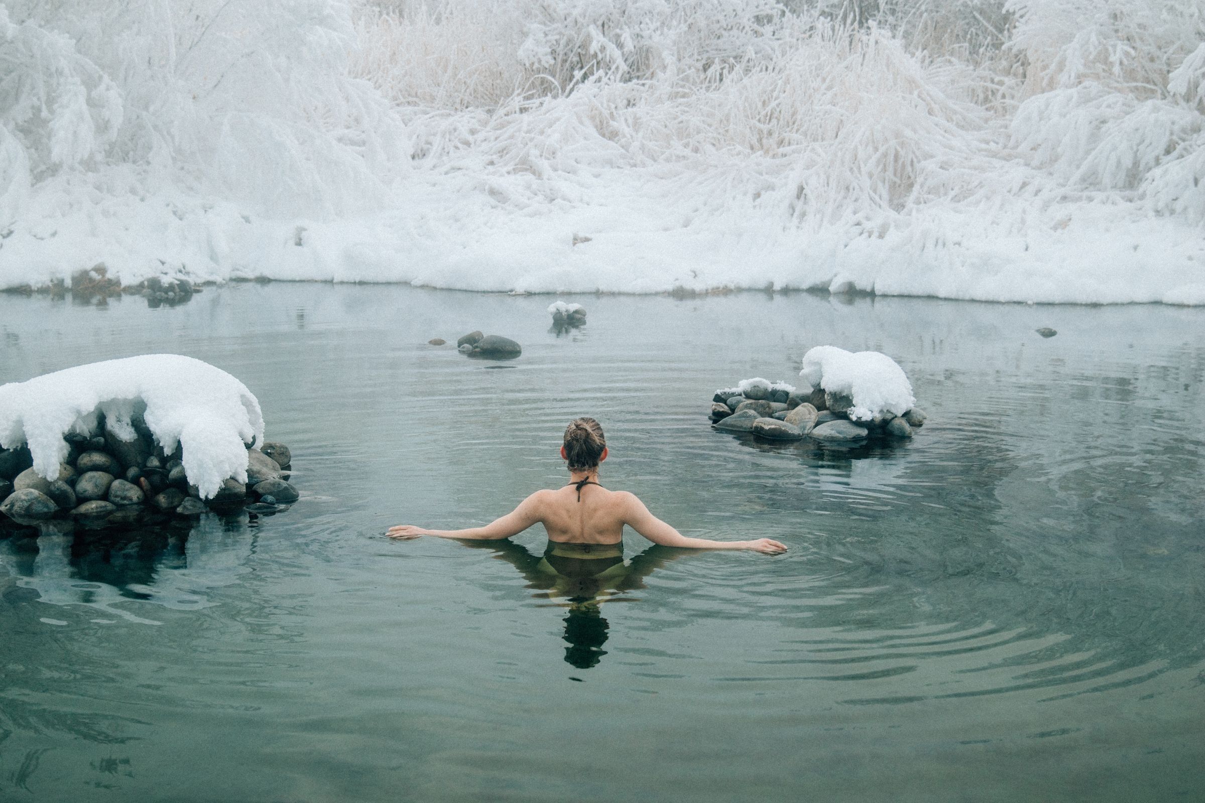 A bare-backed woman with her arms outstretched in a dark, cold pool beside snow-covered rocks and frost-coated grasses.