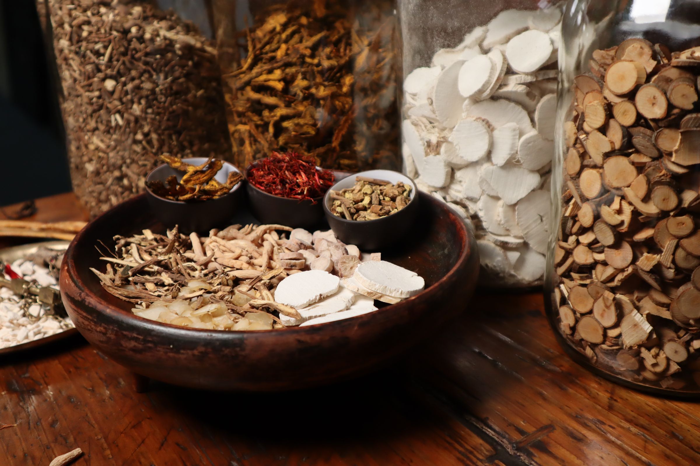Dried Chinese herbal roots and slices in a wooden bowl and glass jars on a wooden table.