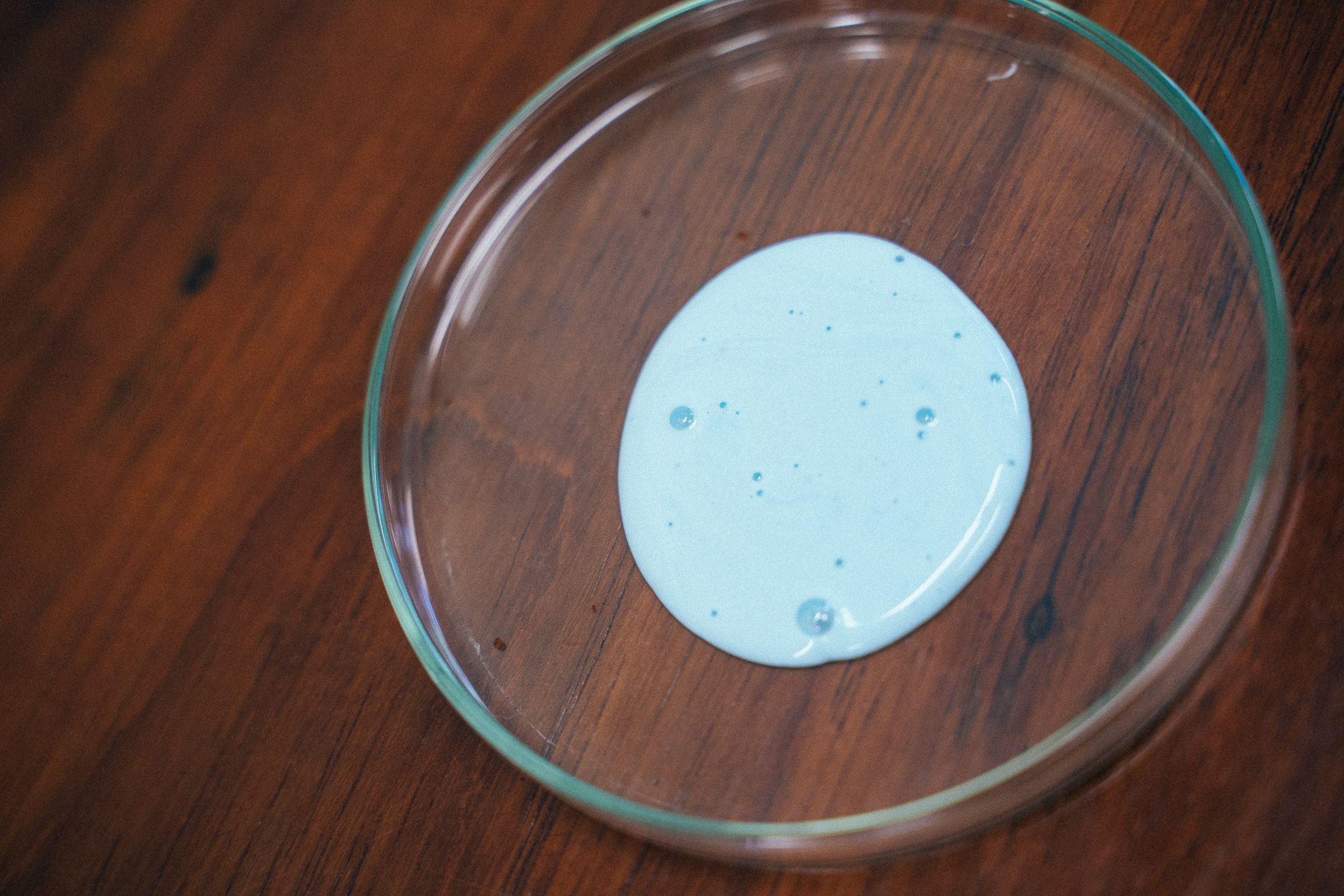 A pale blue liquid spread in a glass petri dish on a wooden table.