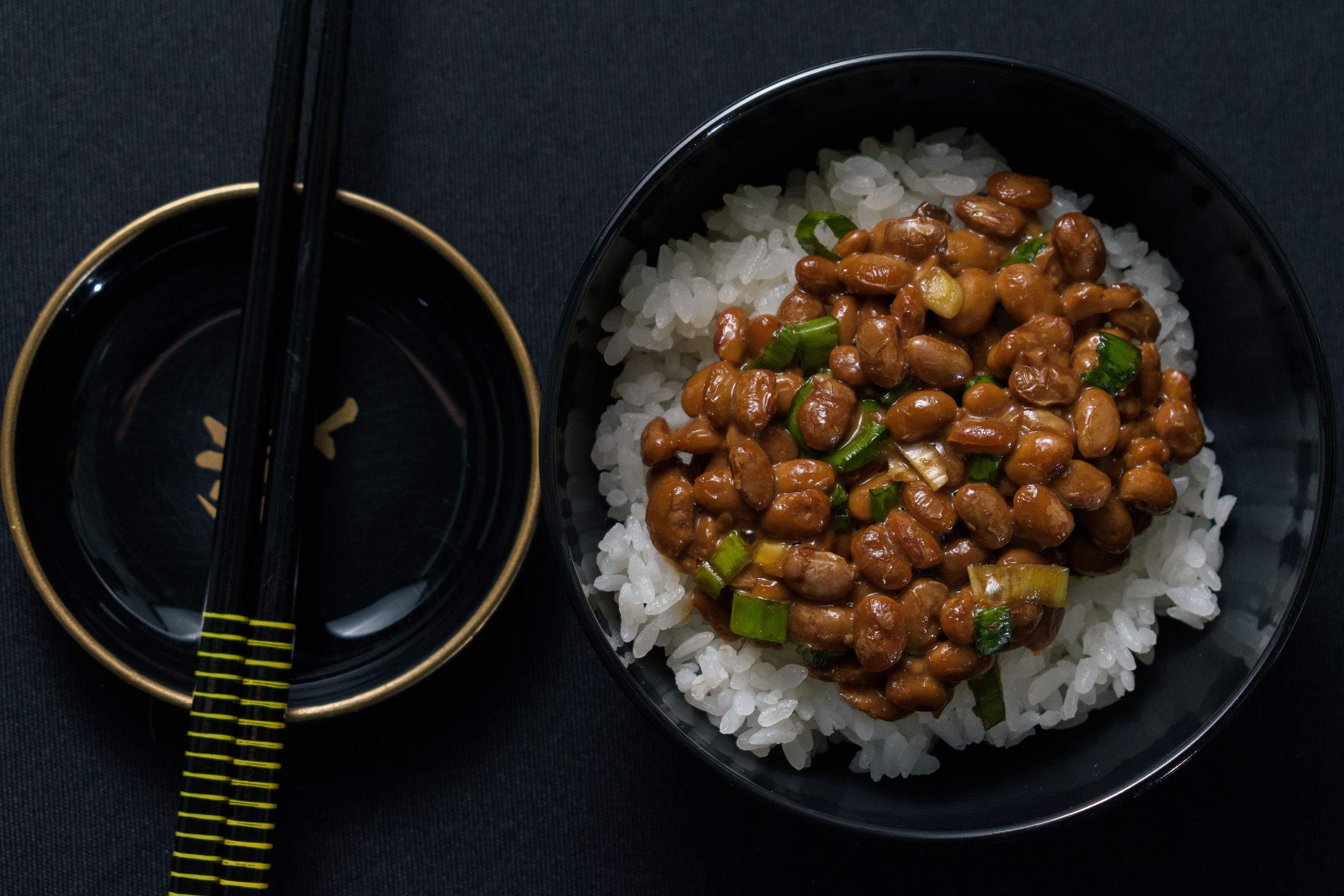 Natto fermented soybeans with green onion over white rice in a black bowl, with chopsticks beside it.