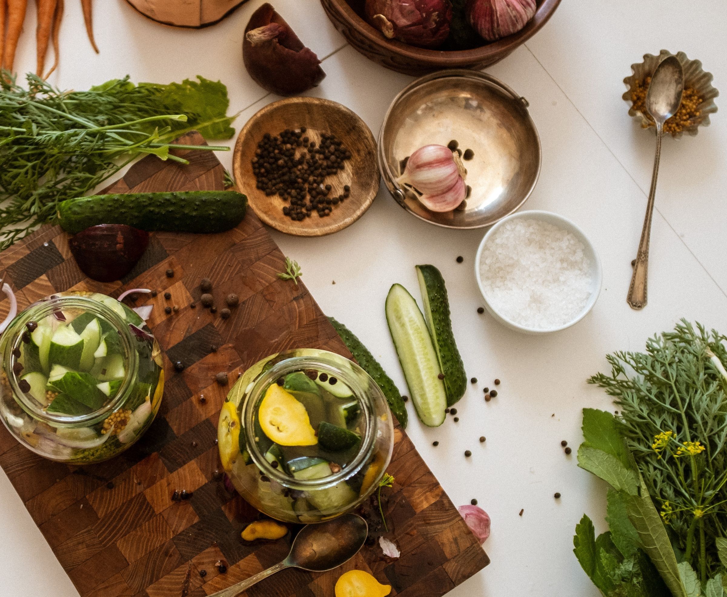 Two glass jars of pickled vegetables with dill and garlic on a wooden board beside fresh cucumbers and herbs.