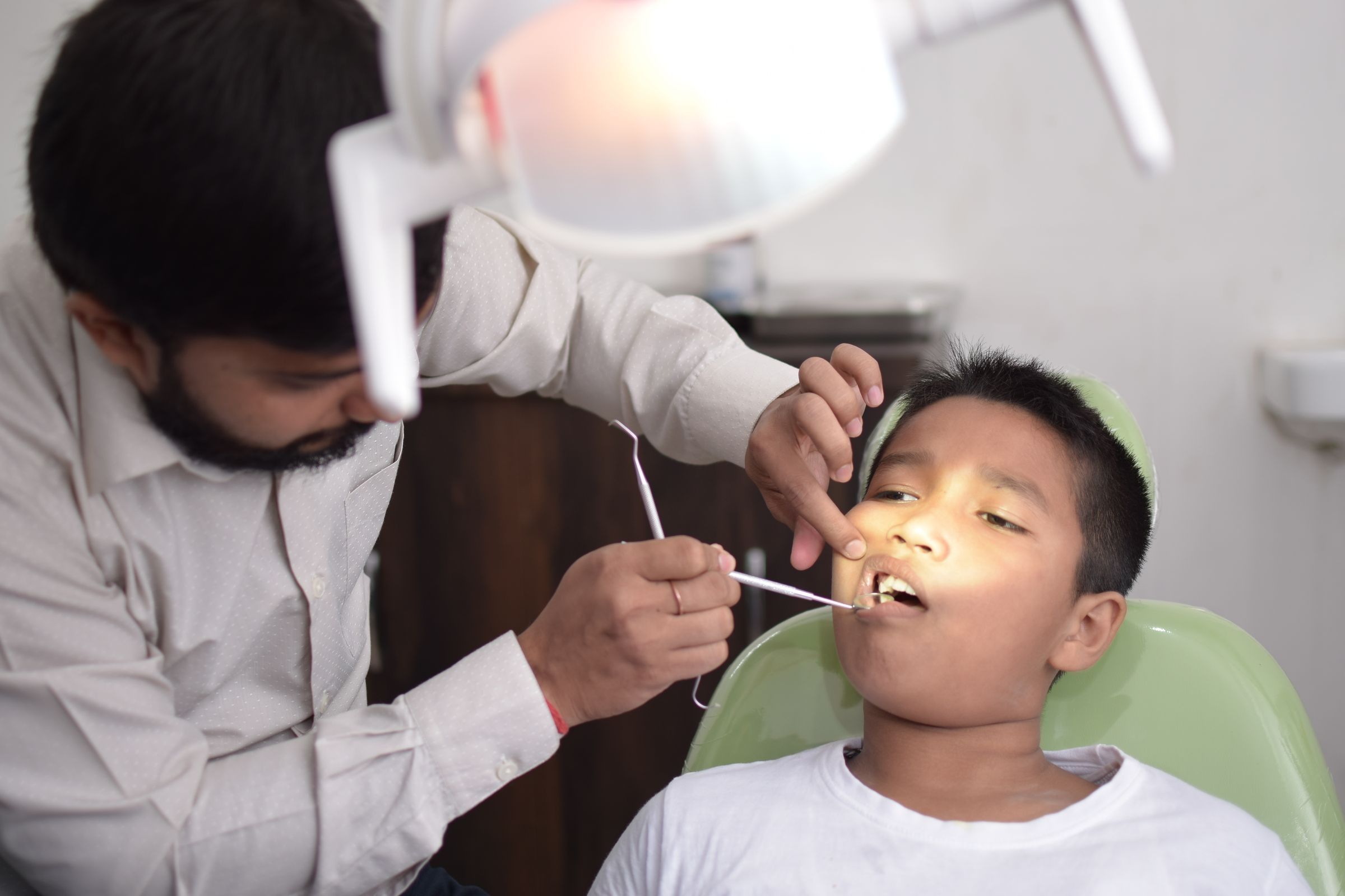 A dentist in a white shirt uses a dental mirror and probe on a young boy seated in a green dental chair, under a bright overhead dental lamp.