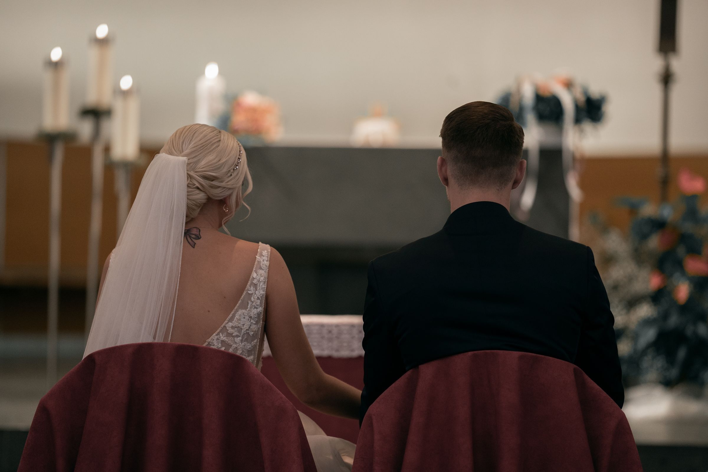 A bride in a veil and lace gown and a groom in a dark suit seen from behind, seated in wooden church pews facing an altar with candles and flowers.