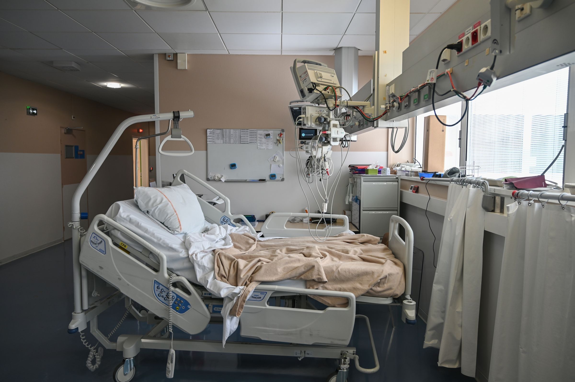An empty hospital room with a hospital bed, monitoring equipment on a wall-mounted arm, and a whiteboard.
