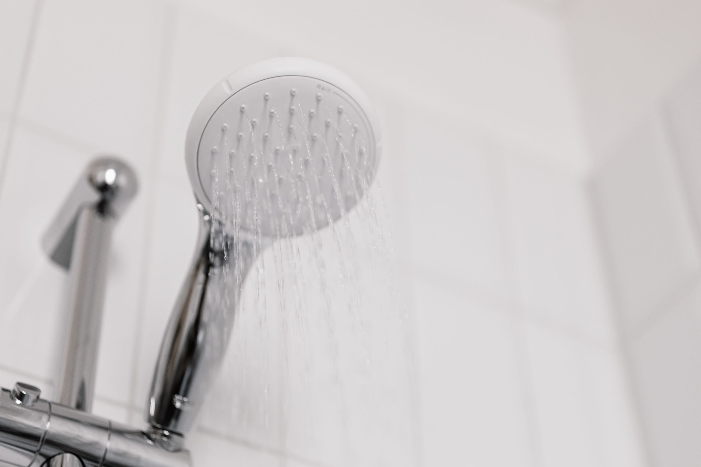 A chrome shower head with water streaming from it against white tiles.