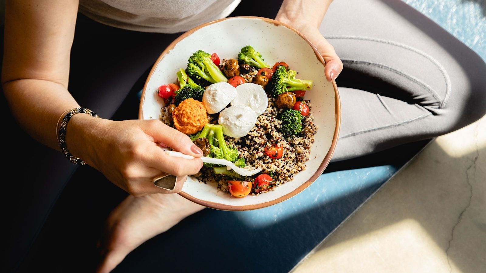 A woman seated on a yoga mat holding a bowl of quinoa, broccoli, tomatoes and poached eggs, seen from above