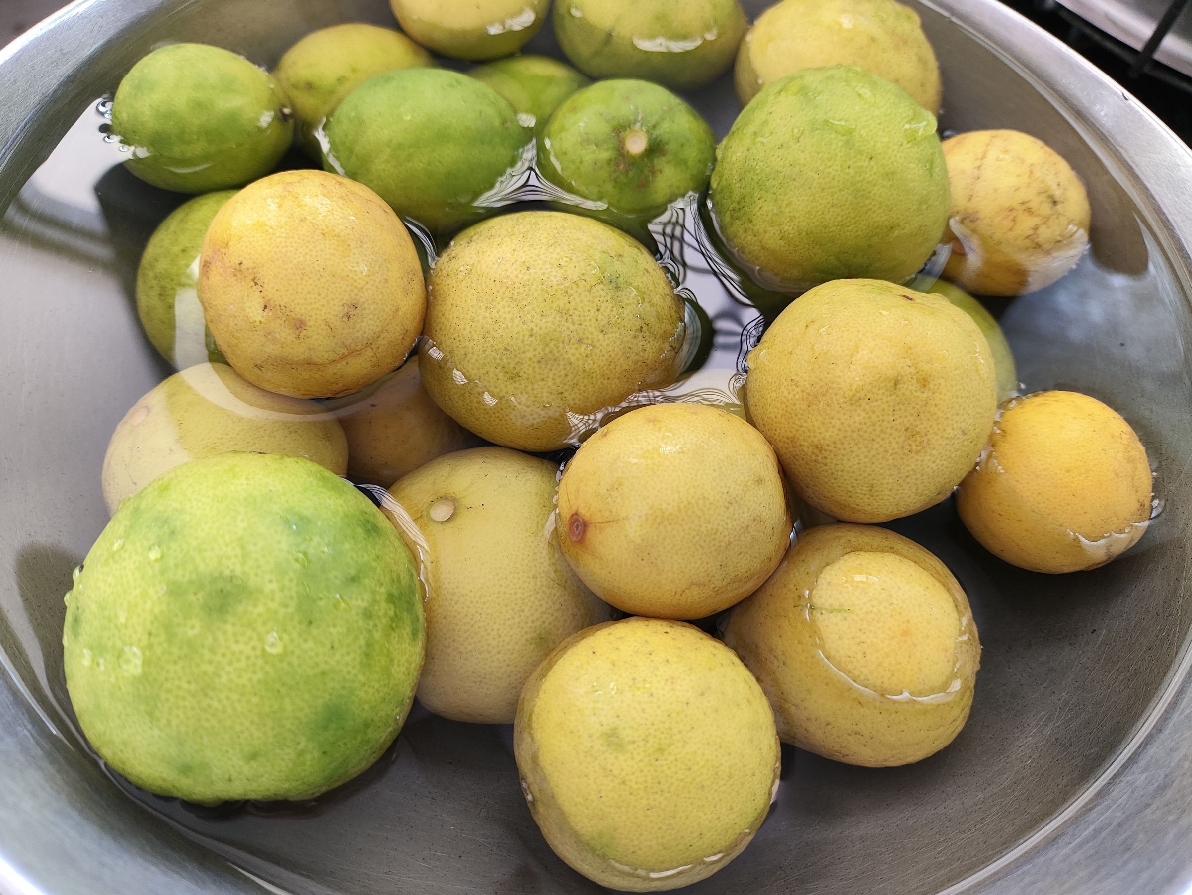 Many green and yellow citrus fruits submerged in water in a metal bowl.