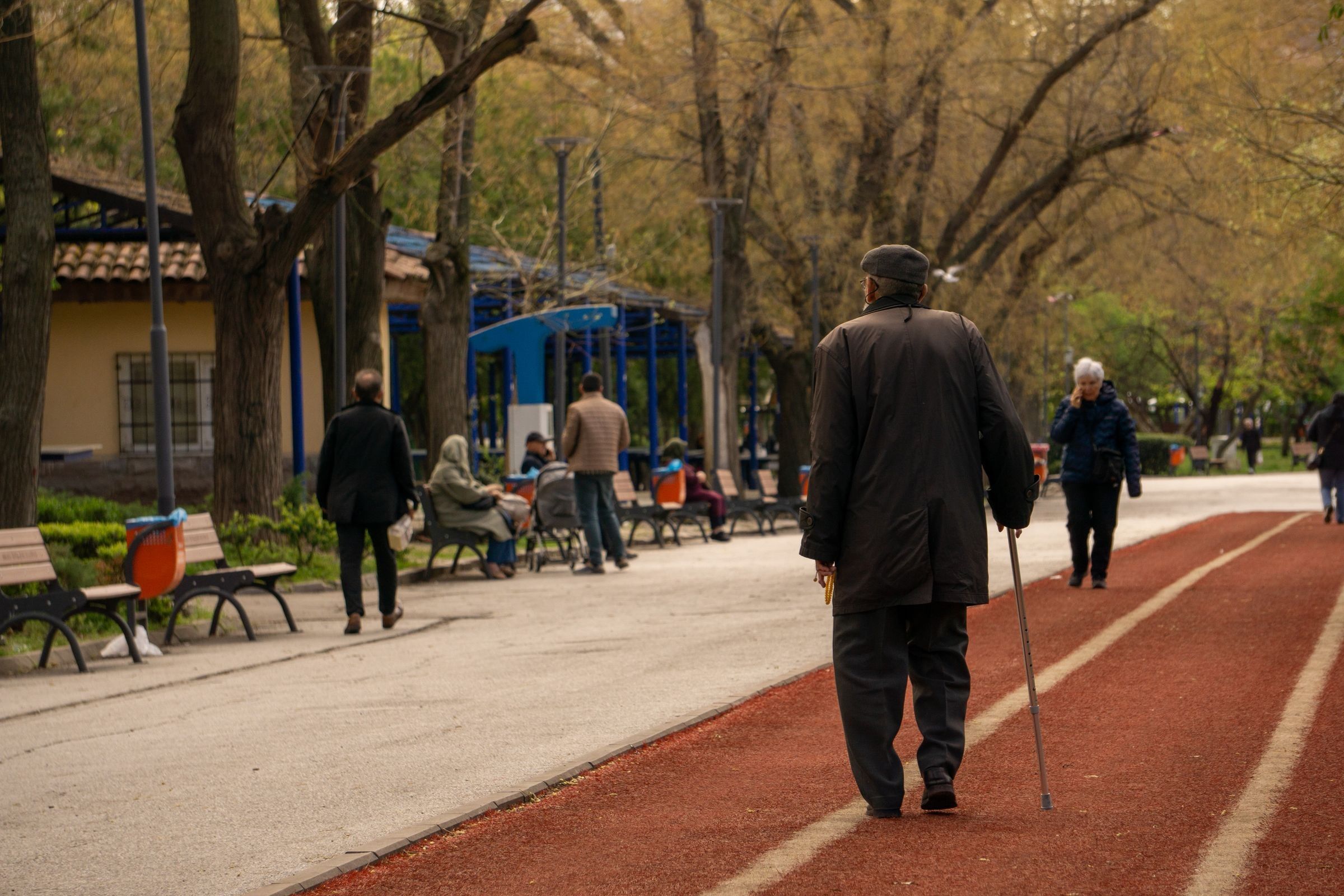 An older man with a walking cane walks on a red running track beside a paved park path, with trees and benches in the background under daylight.