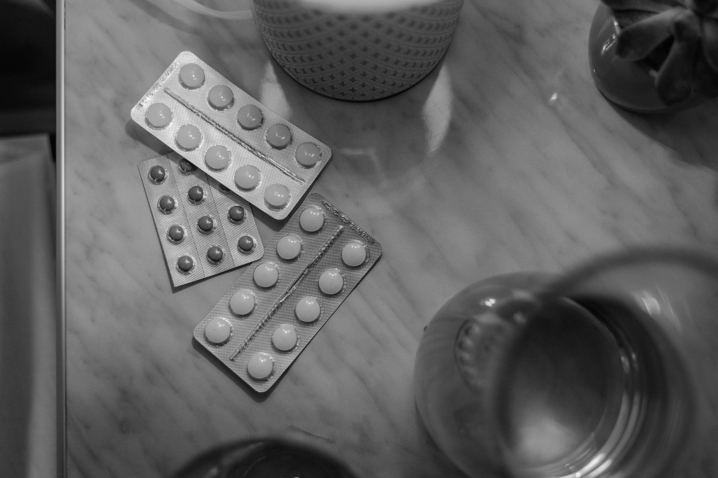 Black and white photo of blister packs of pills on a marble table with a glass and a cup nearby.
