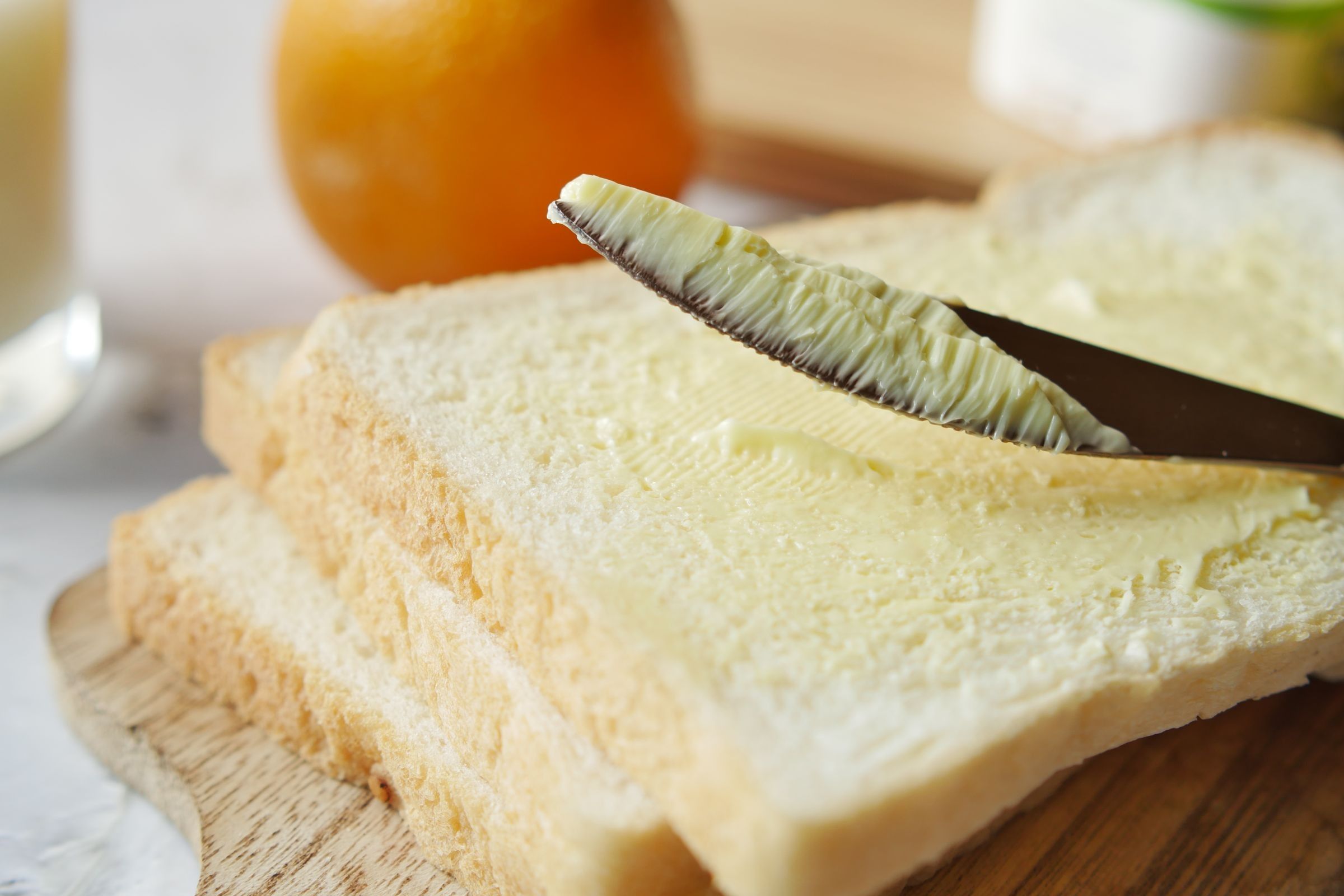 A knife spreading butter on a slice of white bread on a wooden board, with an orange and a glass in the background.