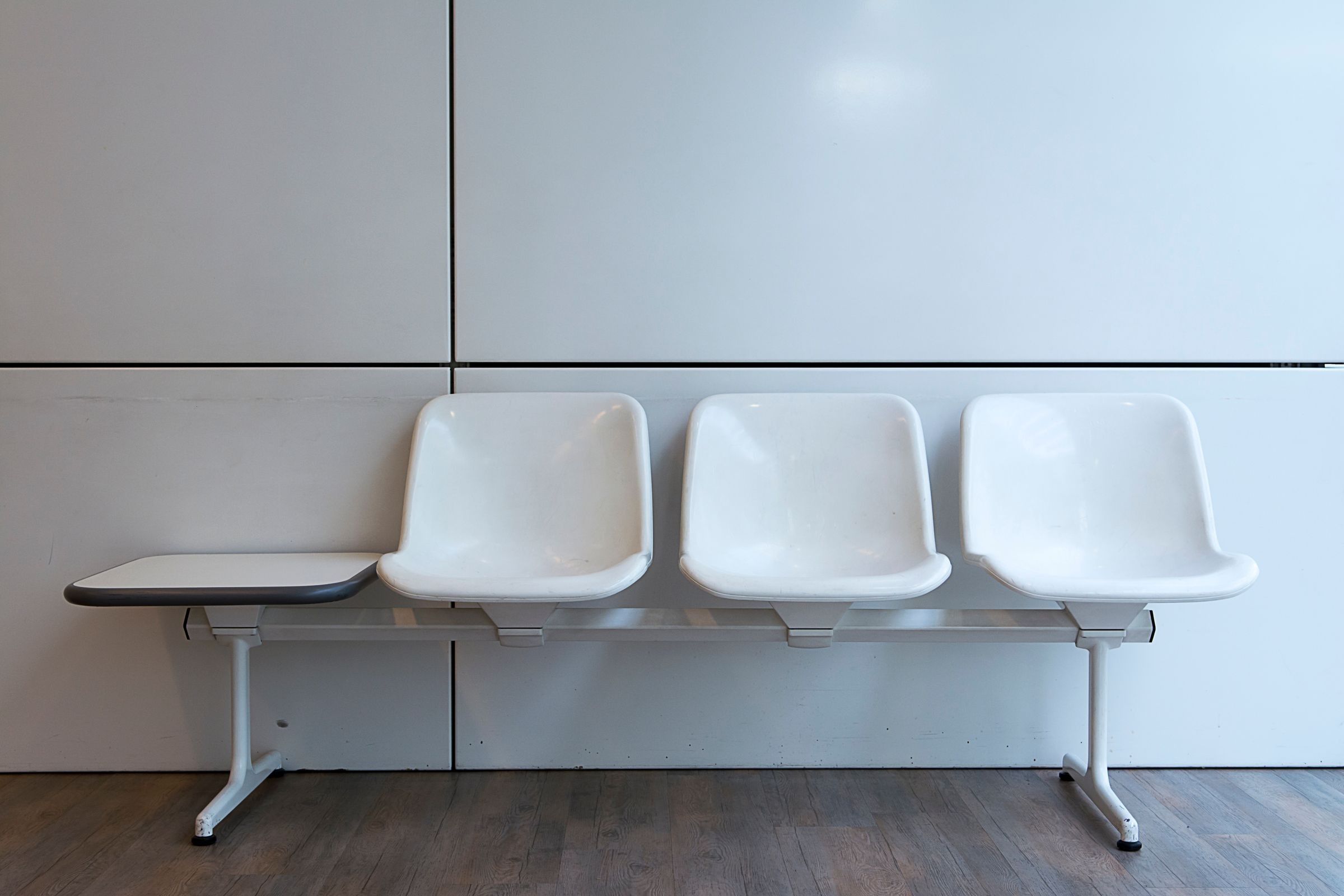Three white plastic chairs and a small attached table against a pale wall in an empty waiting area.