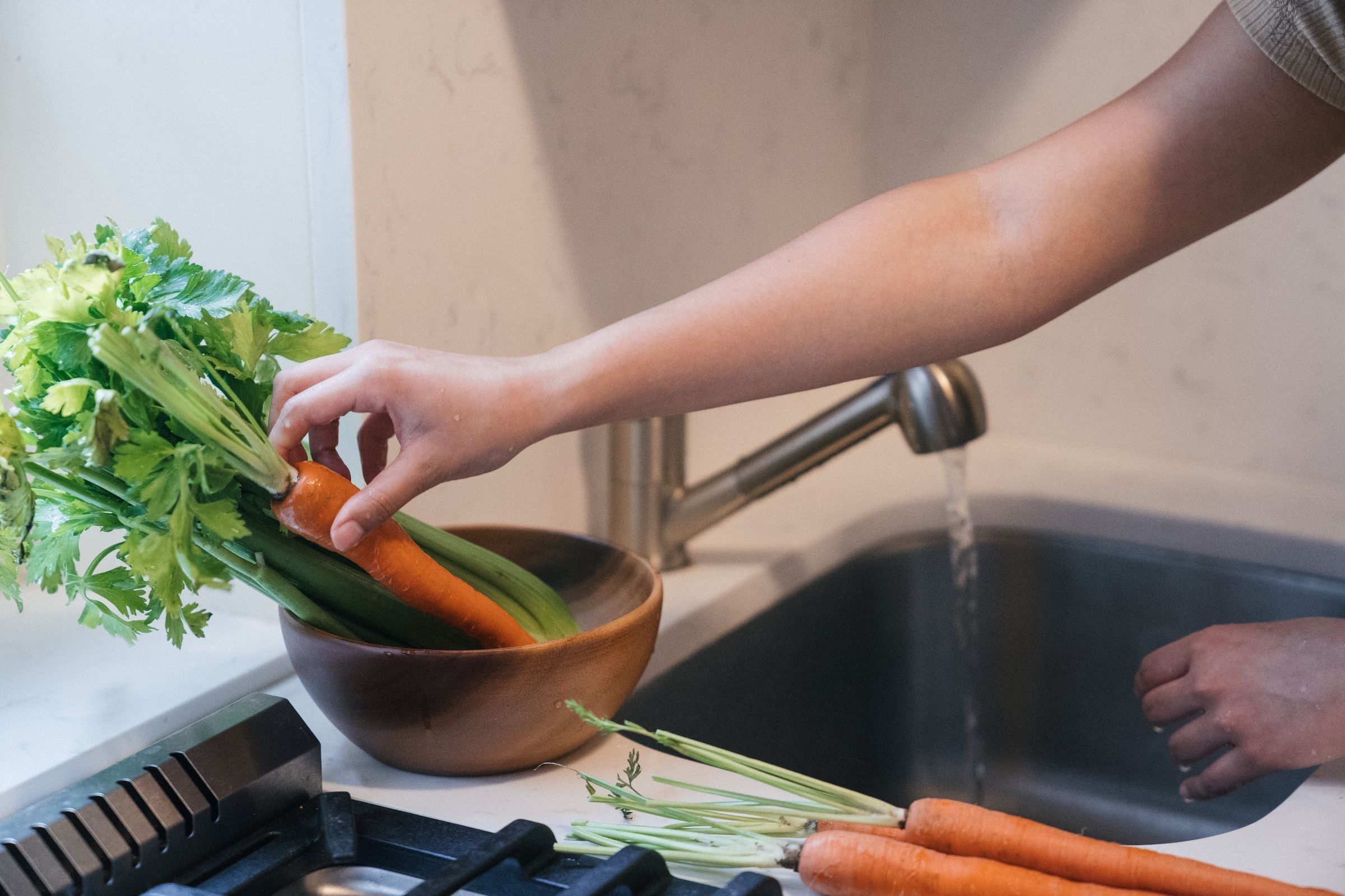 A hand reaching for a carrot in a wooden bowl of green stalks beside a kitchen sink, with carrots and greens on the counter.