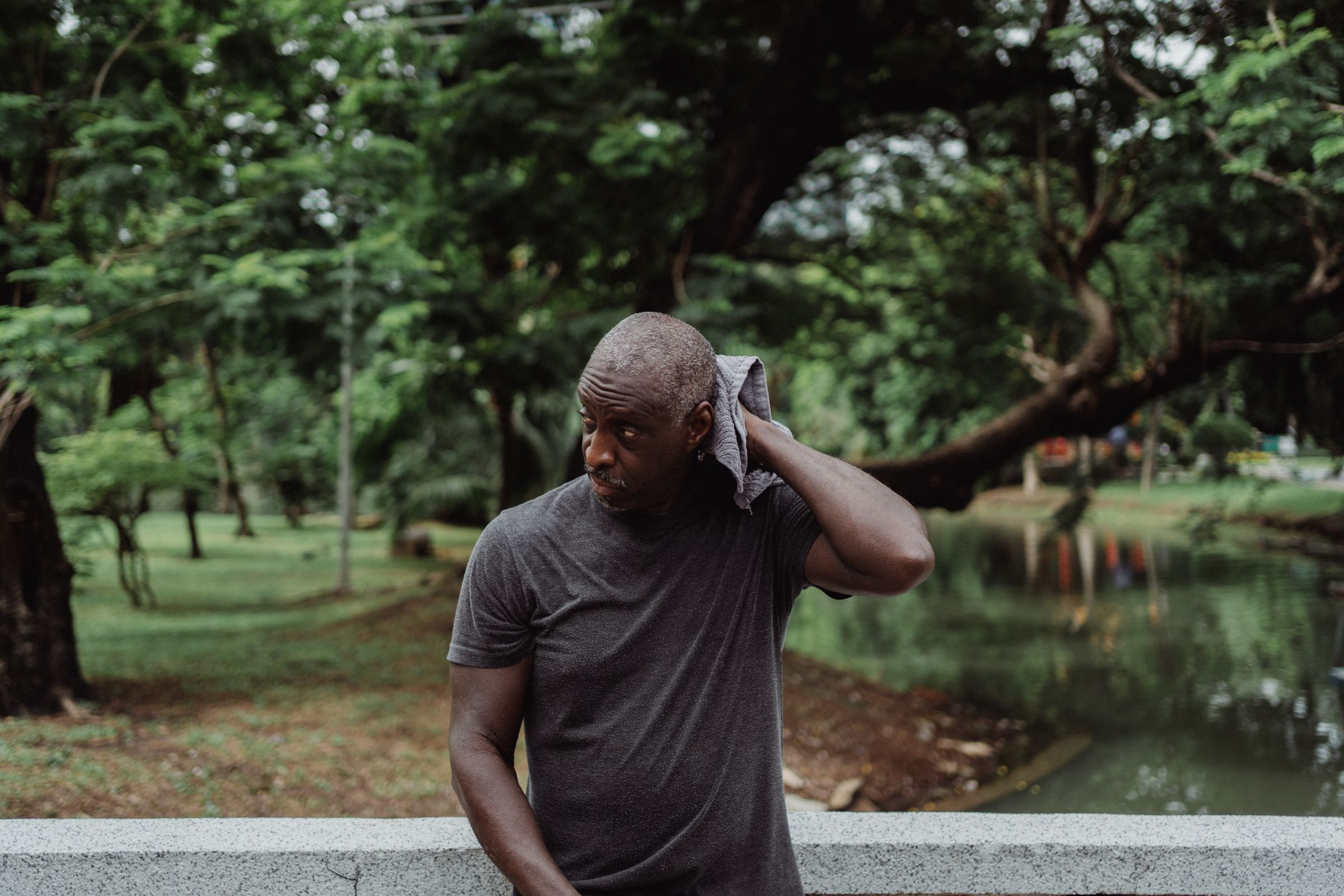 A man in a grey t-shirt wipes his neck with a towel outdoors beside a park pond and green trees.
