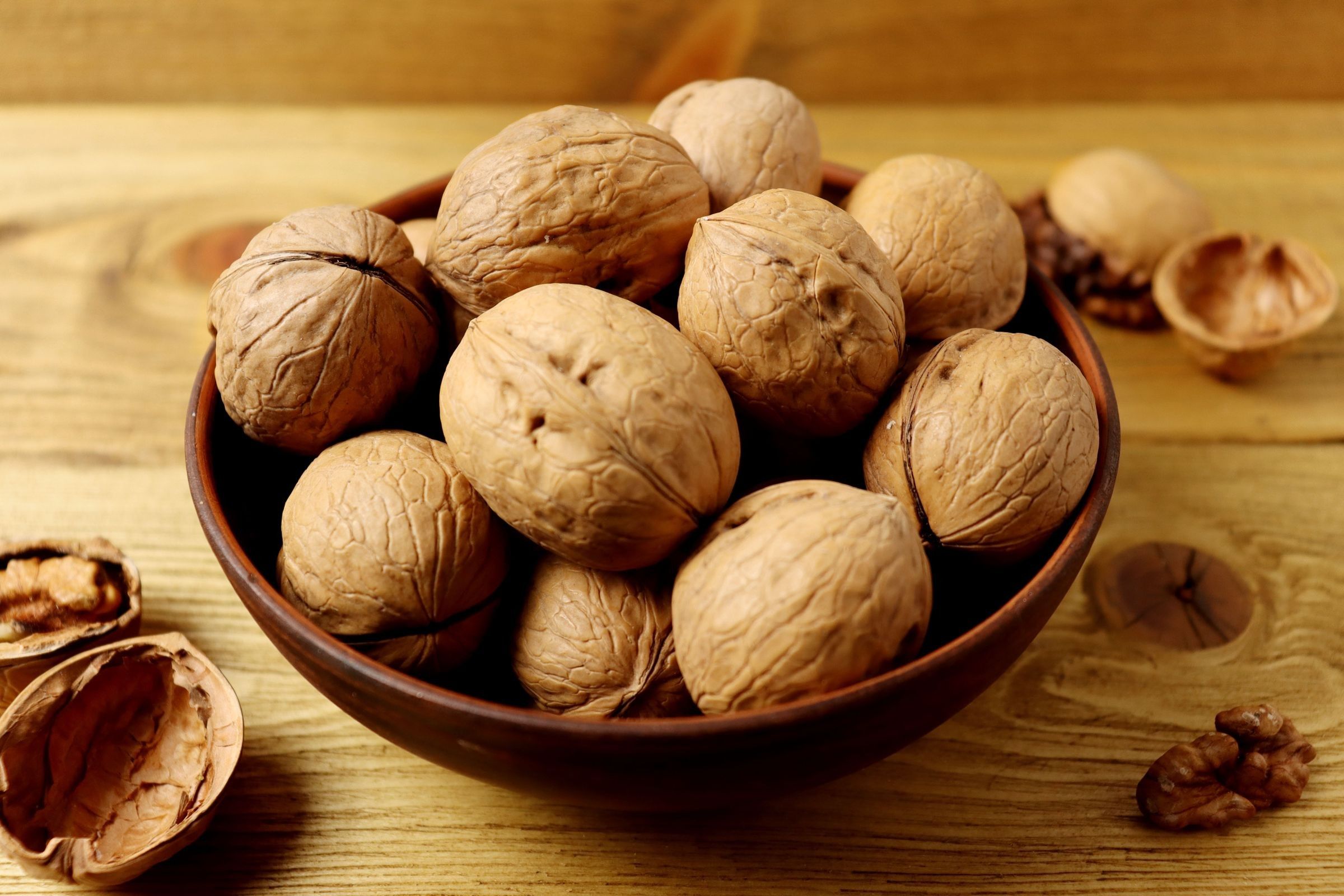 A brown bowl of whole walnuts in shell on a wooden table, with cracked shells nearby.