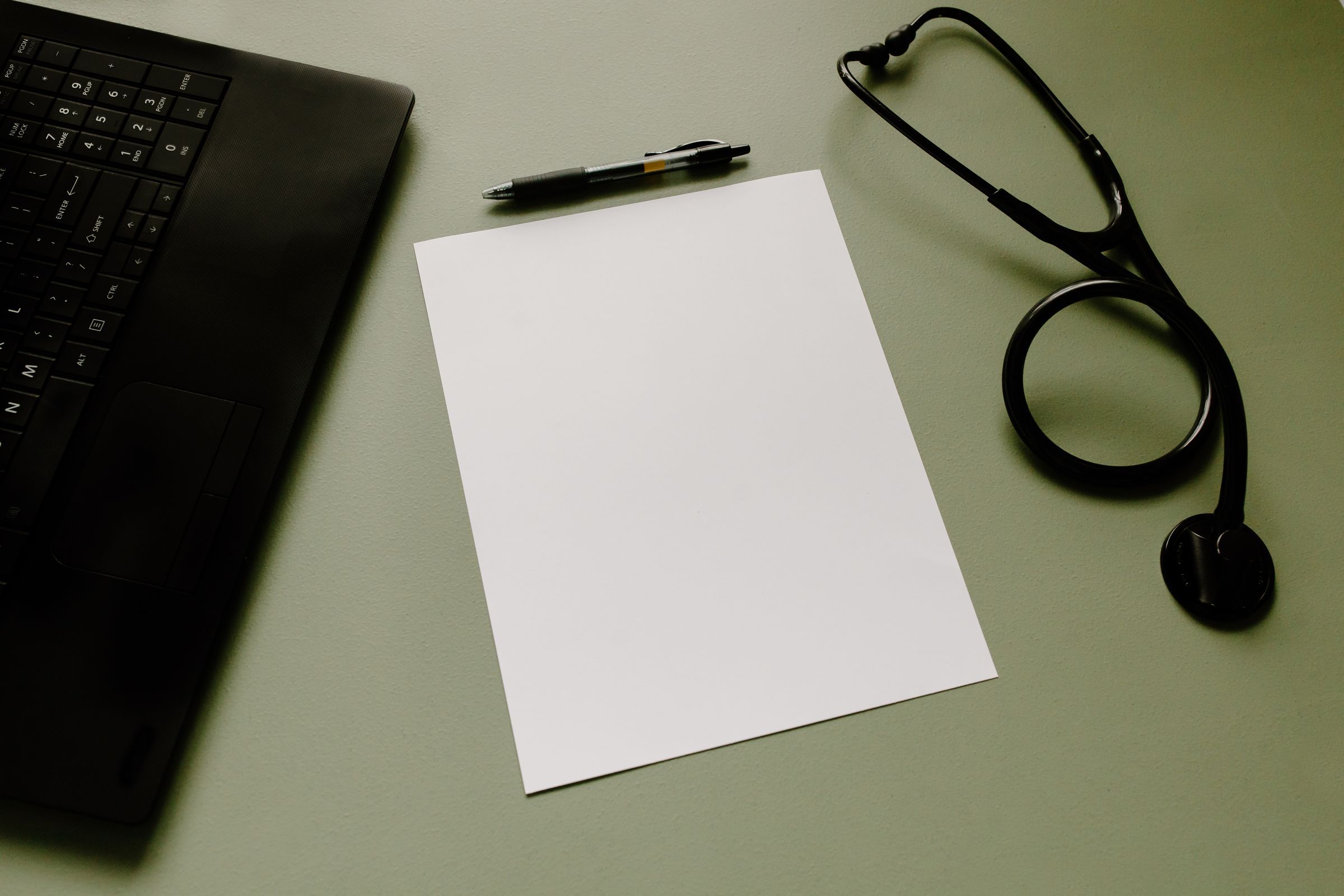 A stethoscope, pen and paper on a desk beside a laptop