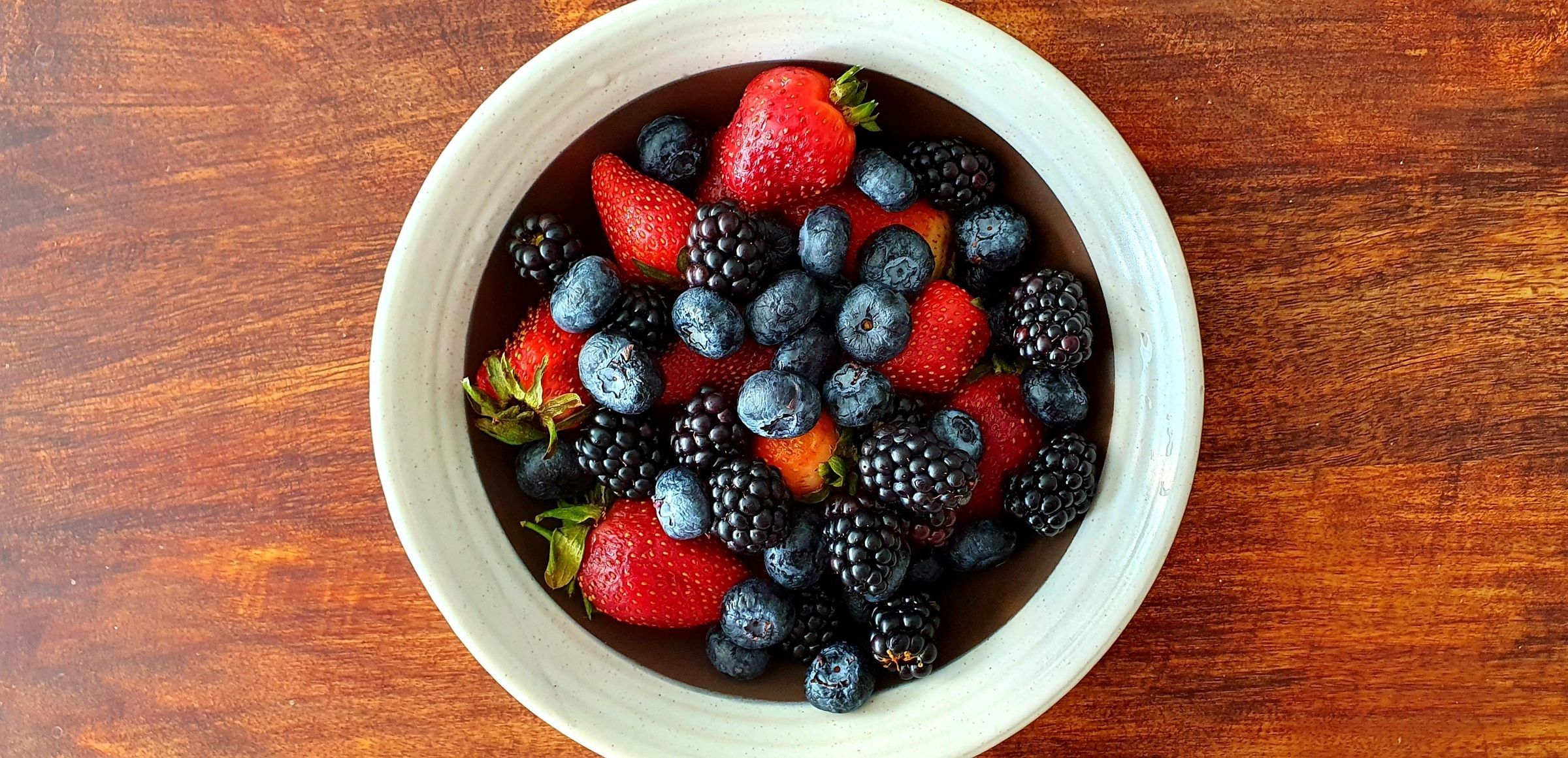 A white ceramic bowl of strawberries, blackberries and blueberries on a wooden table, seen from above.