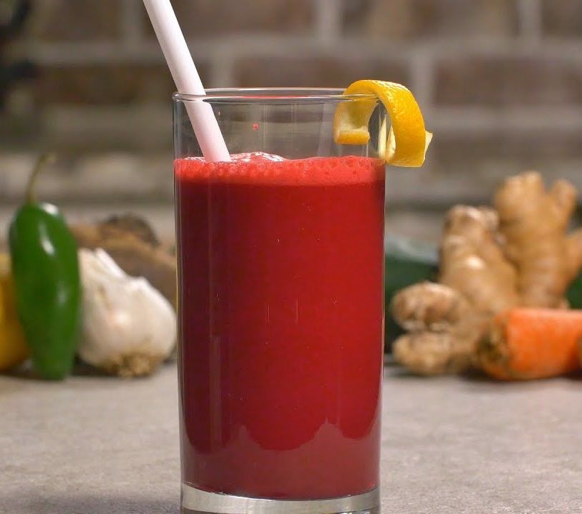 Hot Heart Health Juice in a glass with a lemon twist, with jalapeño, garlic, ginger and carrot behind it