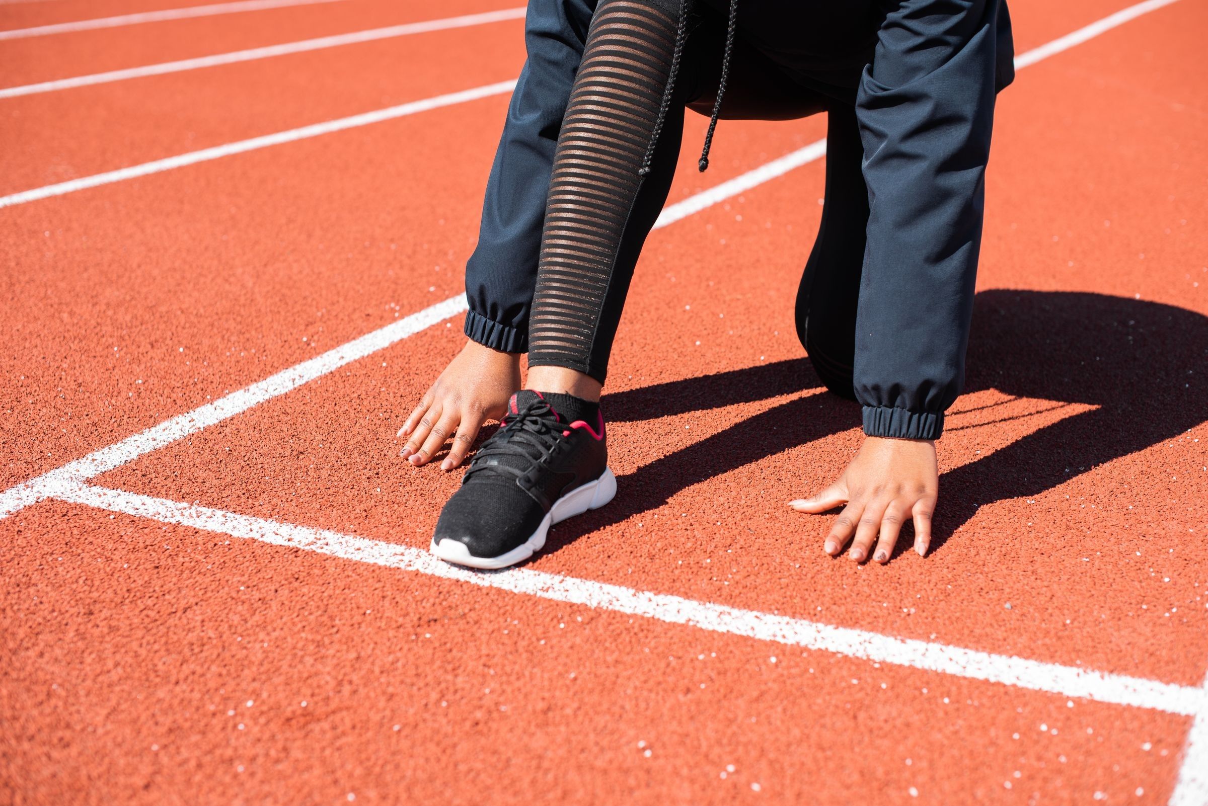 A person in dark athletic clothing and black sneakers crouches in a starting position on an orange running track with white lane lines.