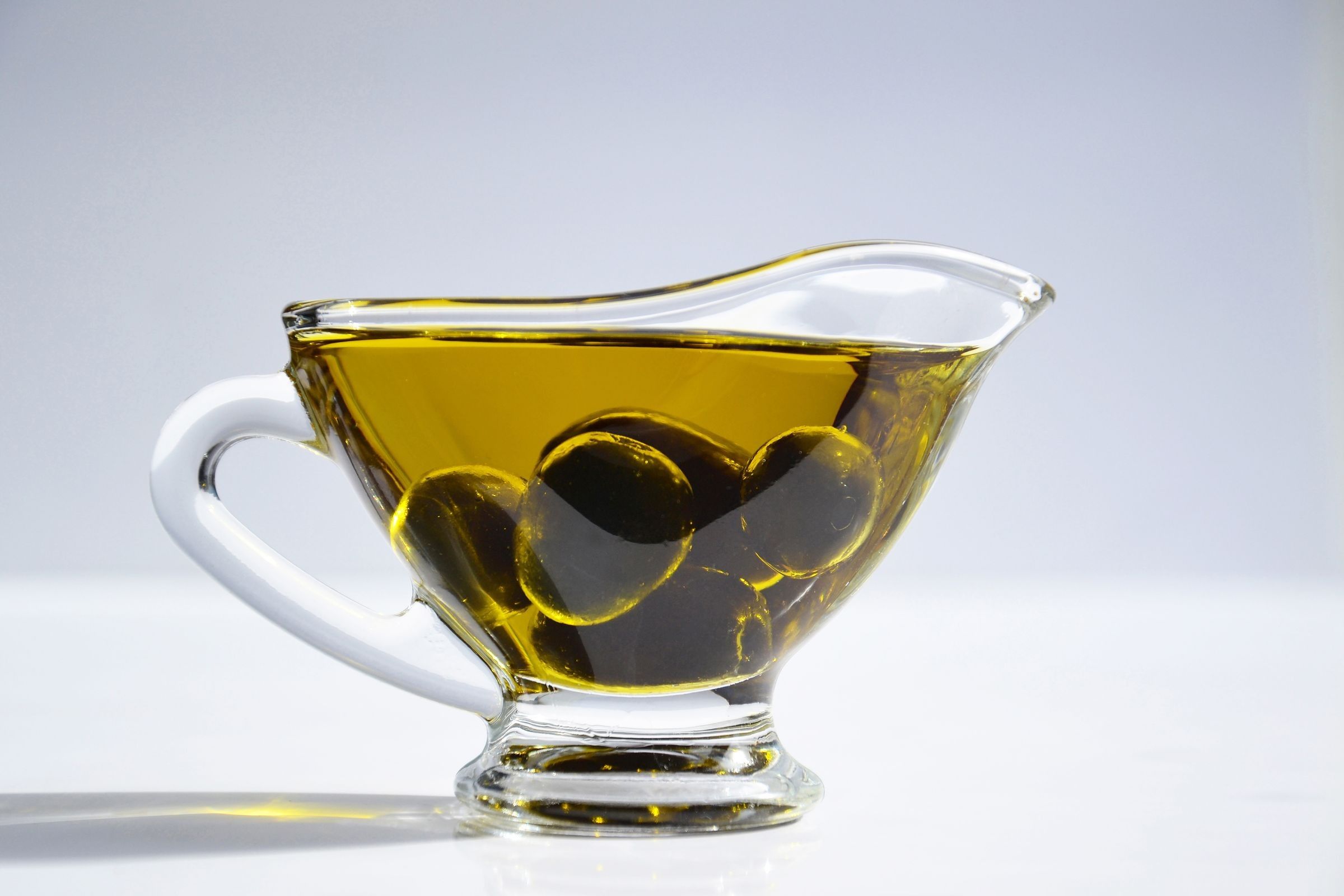 A clear glass gravy boat filled with golden oil and dark olives on a white surface against a plain light background.