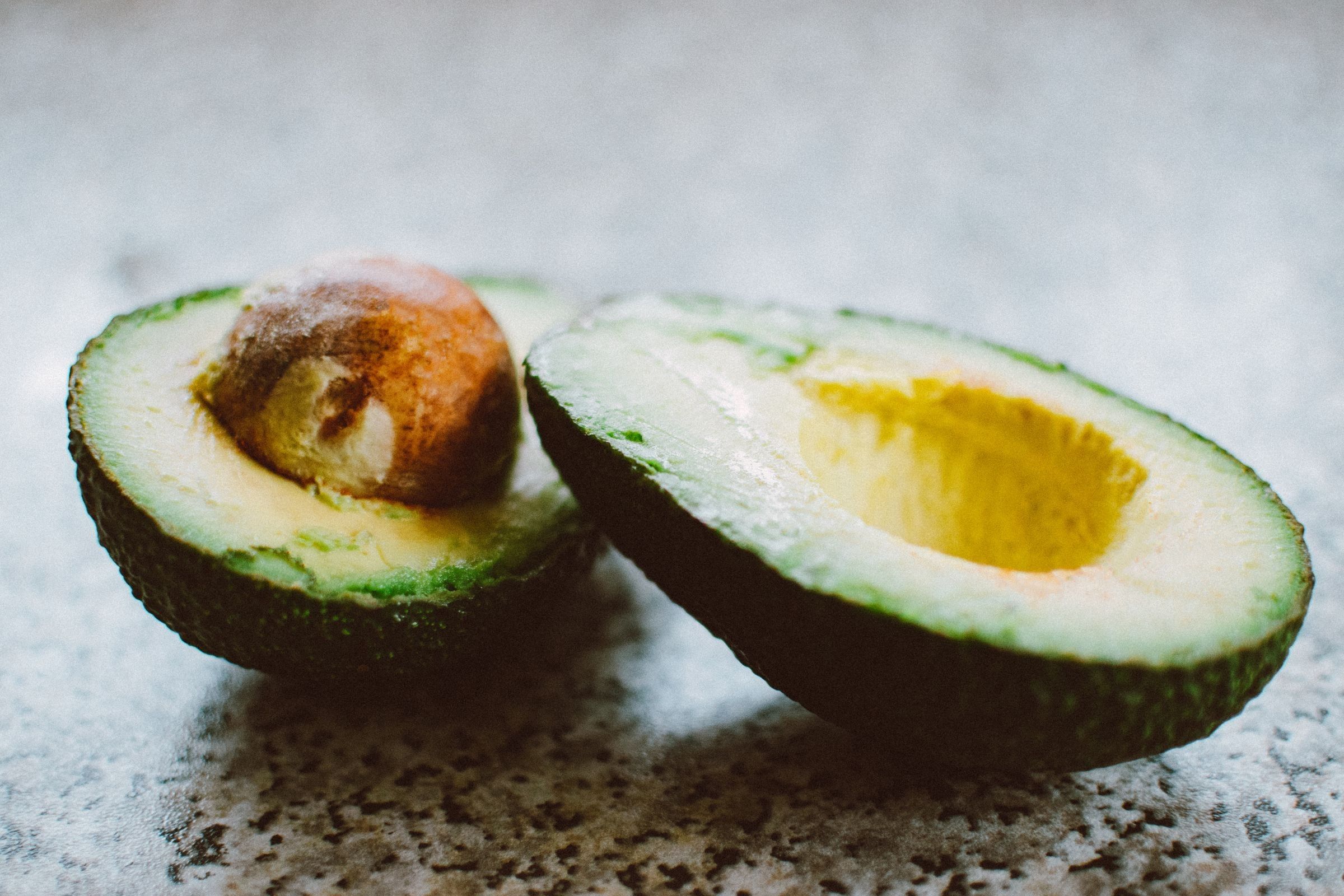 Two halves of a ripe avocado on a grey speckled surface, one showing its pit.