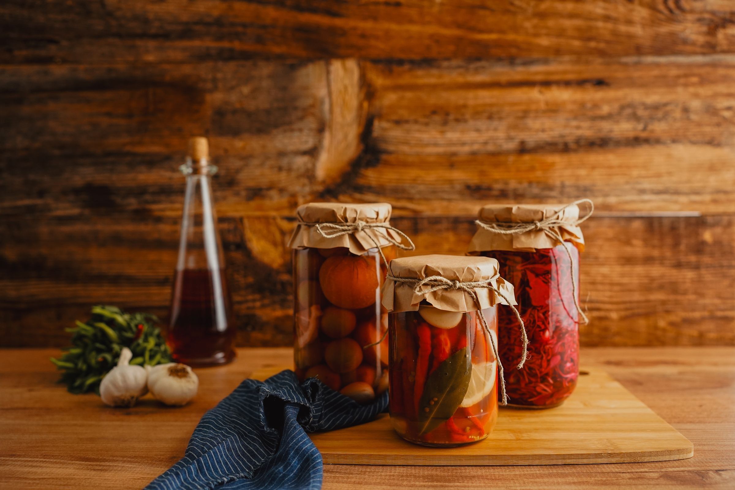 Three glass jars of pickled tomatoes and peppers with bay leaves and garlic on a wooden board, beside a glass bottle of amber liquid.