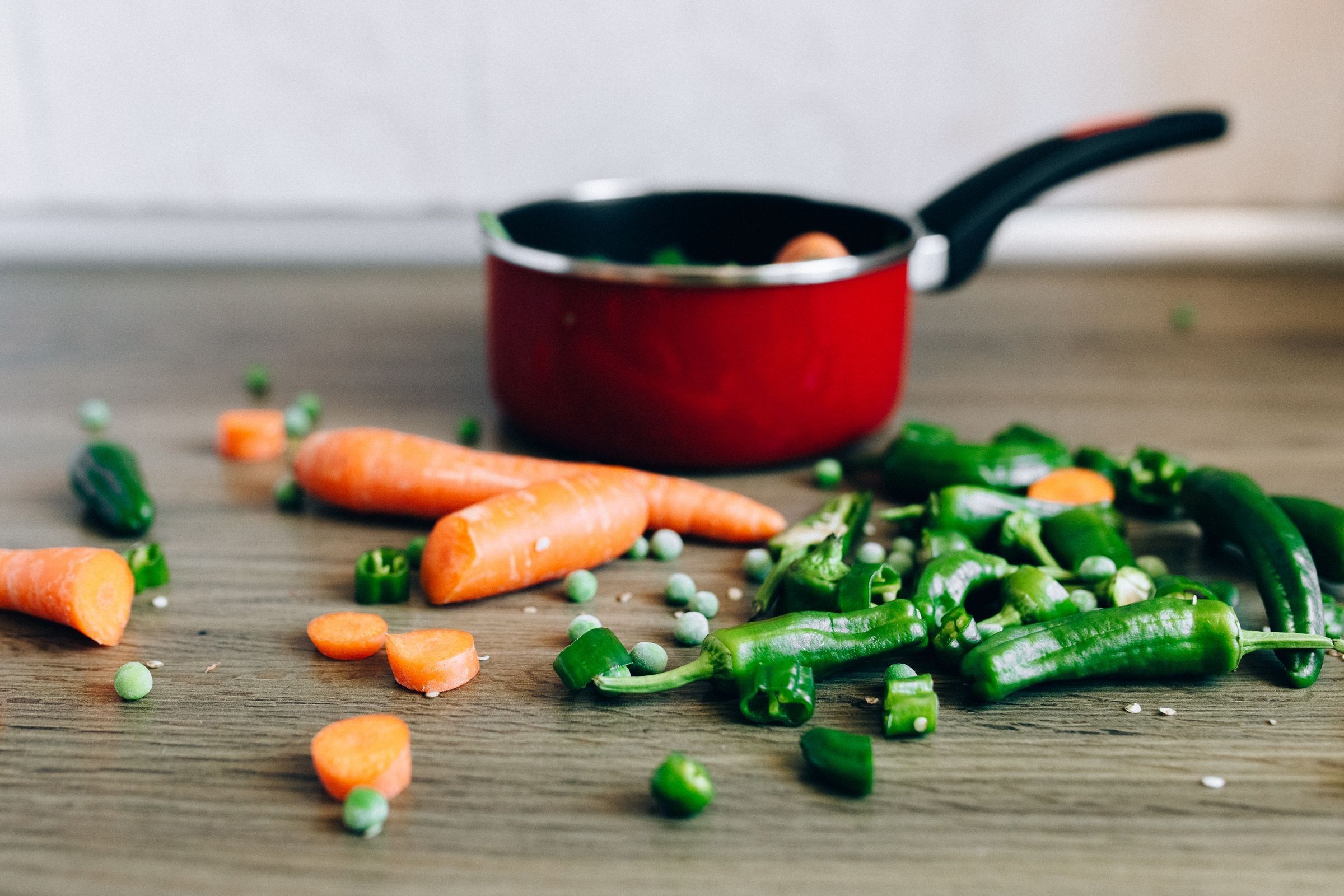 Sliced carrots, green peas and green chili peppers scattered on a wooden table in front of a red saucepan.