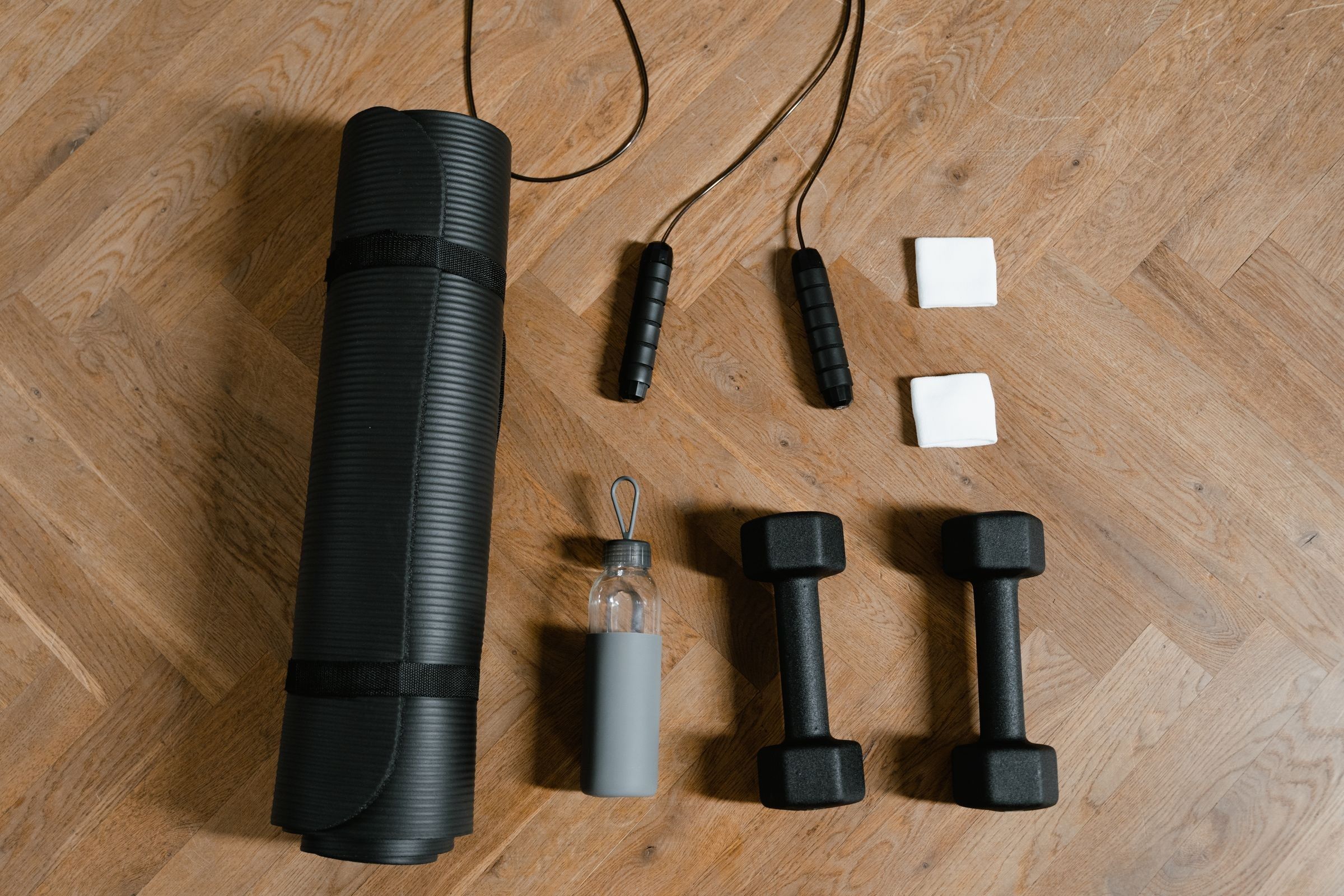 Flat lay of a rolled black exercise mat, two jump ropes, a glass water bottle, two black dumbbells and two white cubes on a wooden floor.