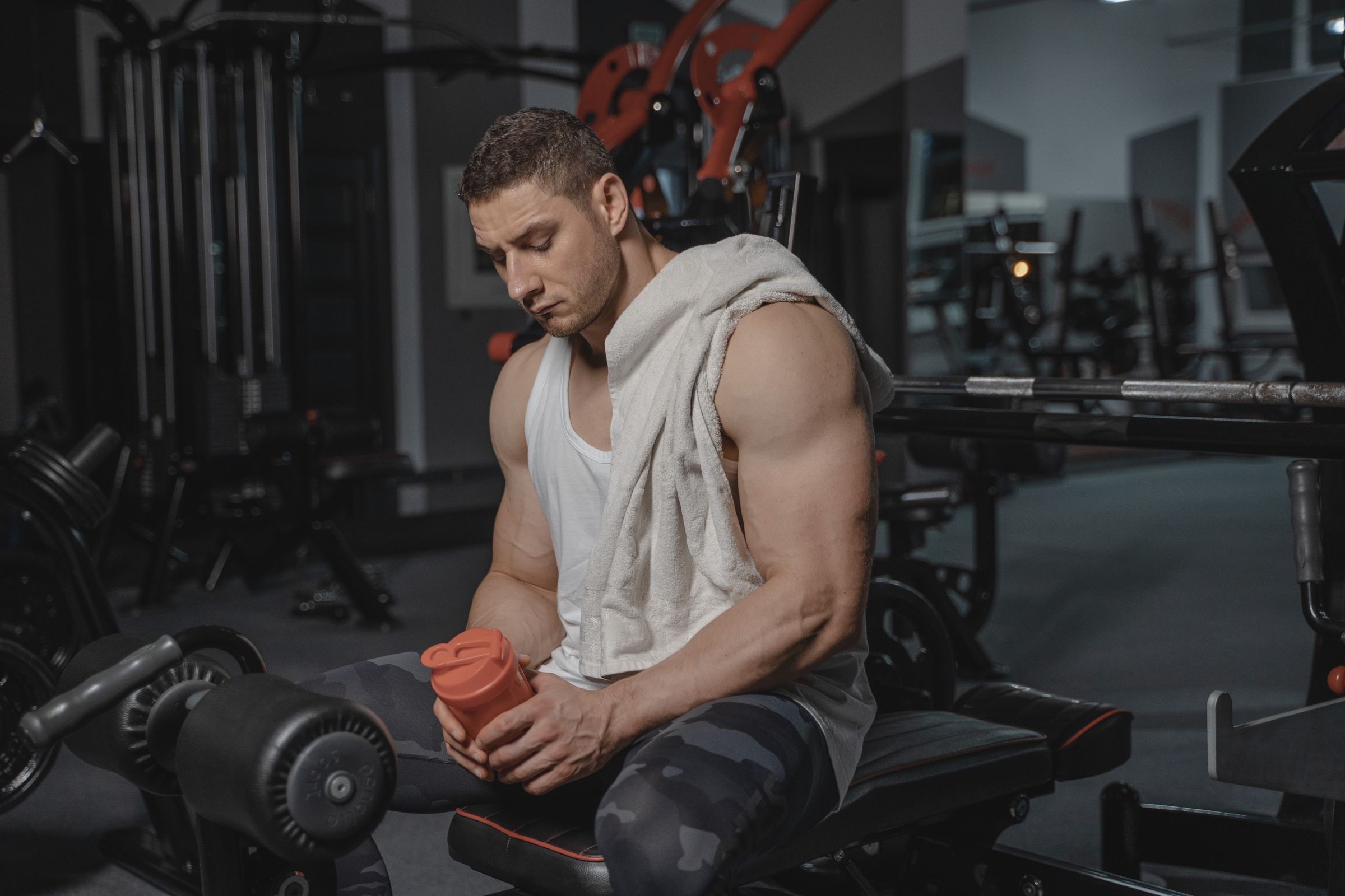 A man with a towel over his shoulder sits on a gym bench holding a red shaker bottle, with weight racks and equipment in the background.