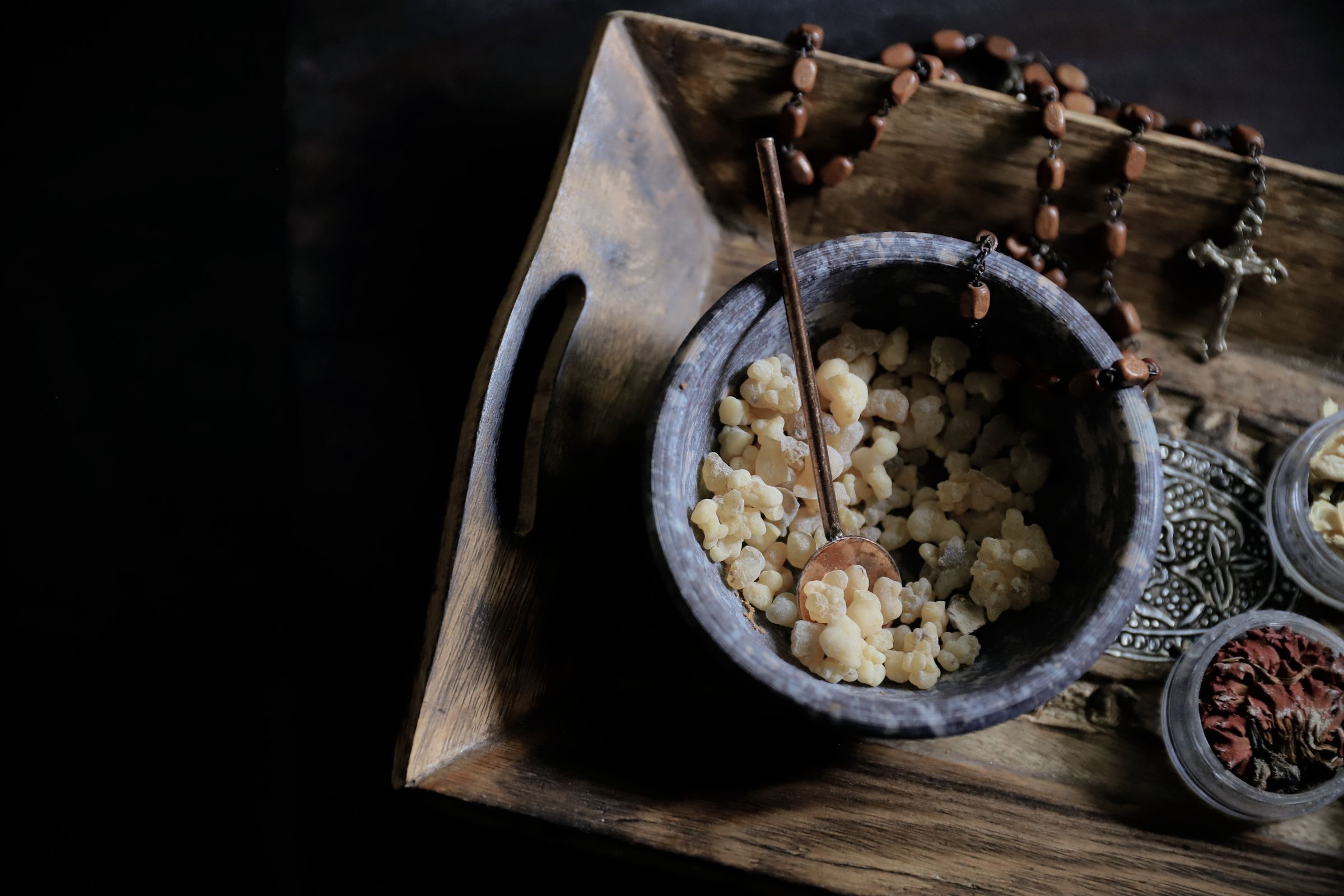 A bowl of pale amber resin pieces on a wooden tray with a wooden spoon, beside a brown bead necklace and small metal and dish items on a dark background.
